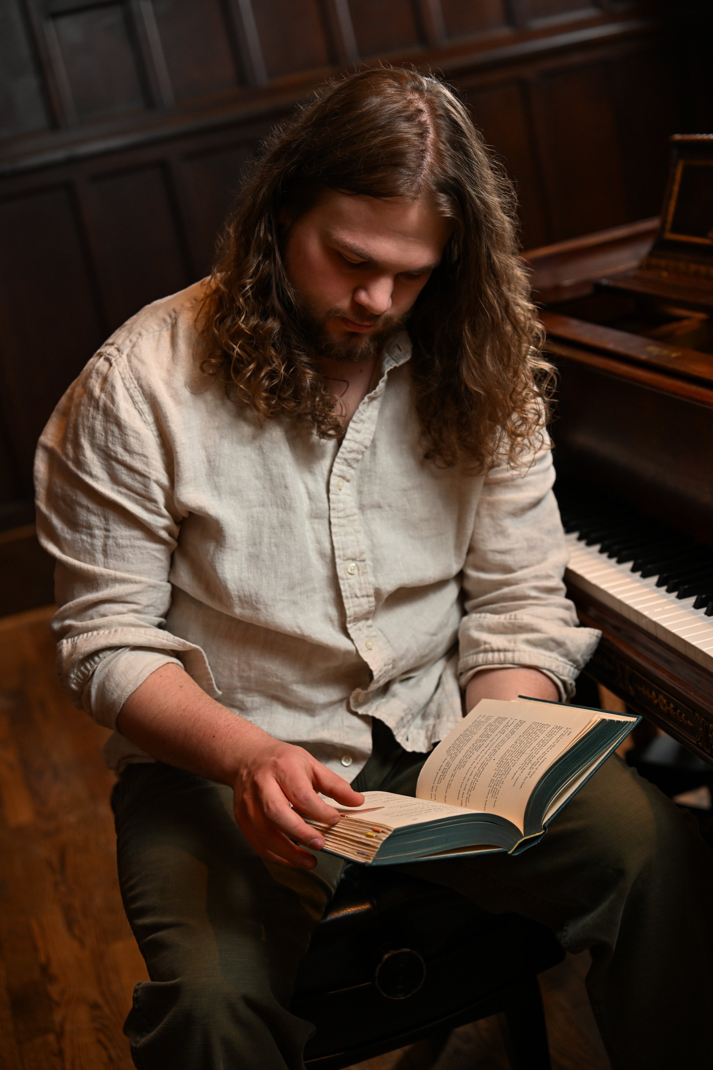 A man with long curly hair and a light-colored shirt sitting at a piano, looking down at an open book in his hands, in a wood-paneled room.
