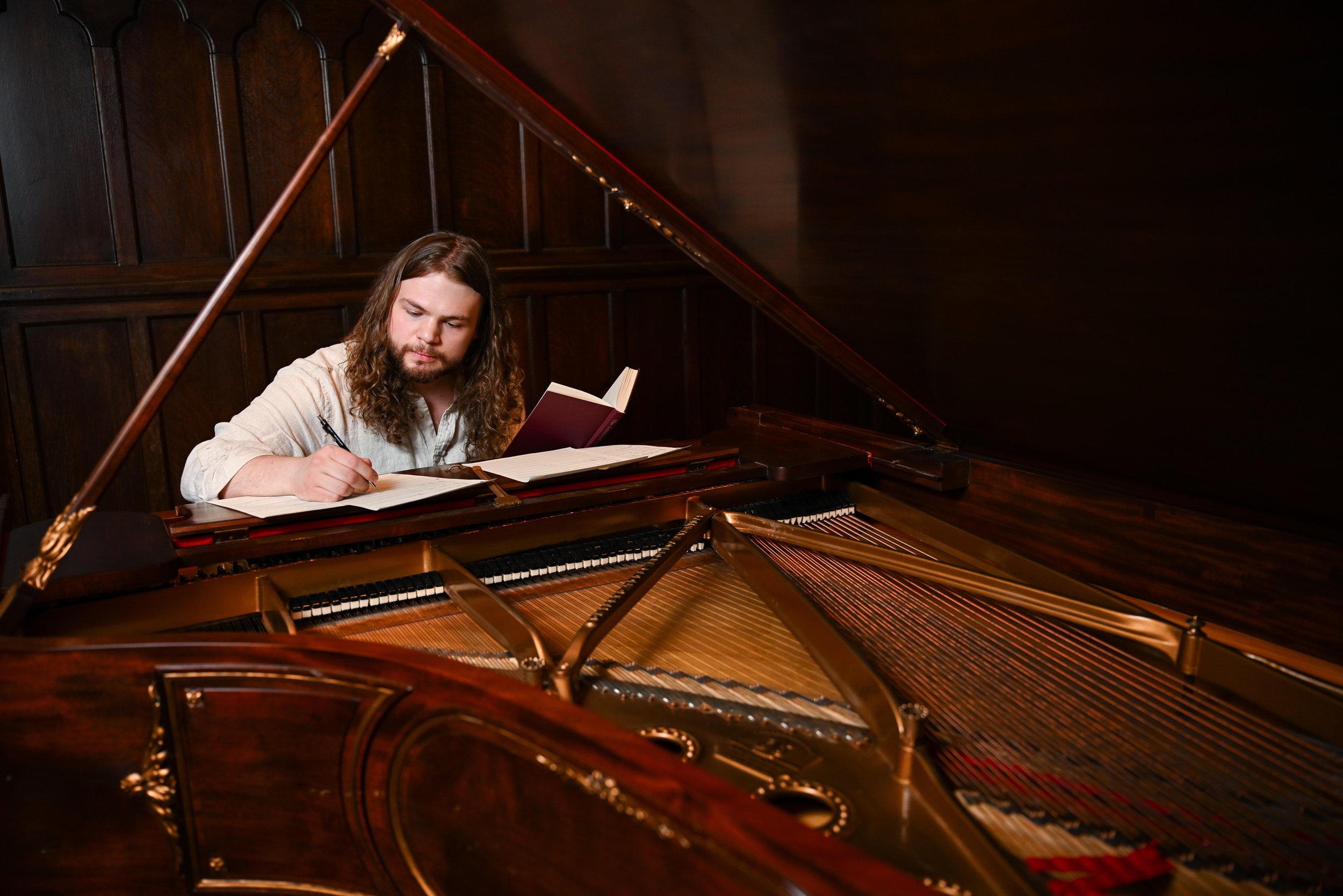 A man with long hair and a beard sitting at a grand piano, writing in a notebook with an open book nearby, inside a room with dark wood paneling.