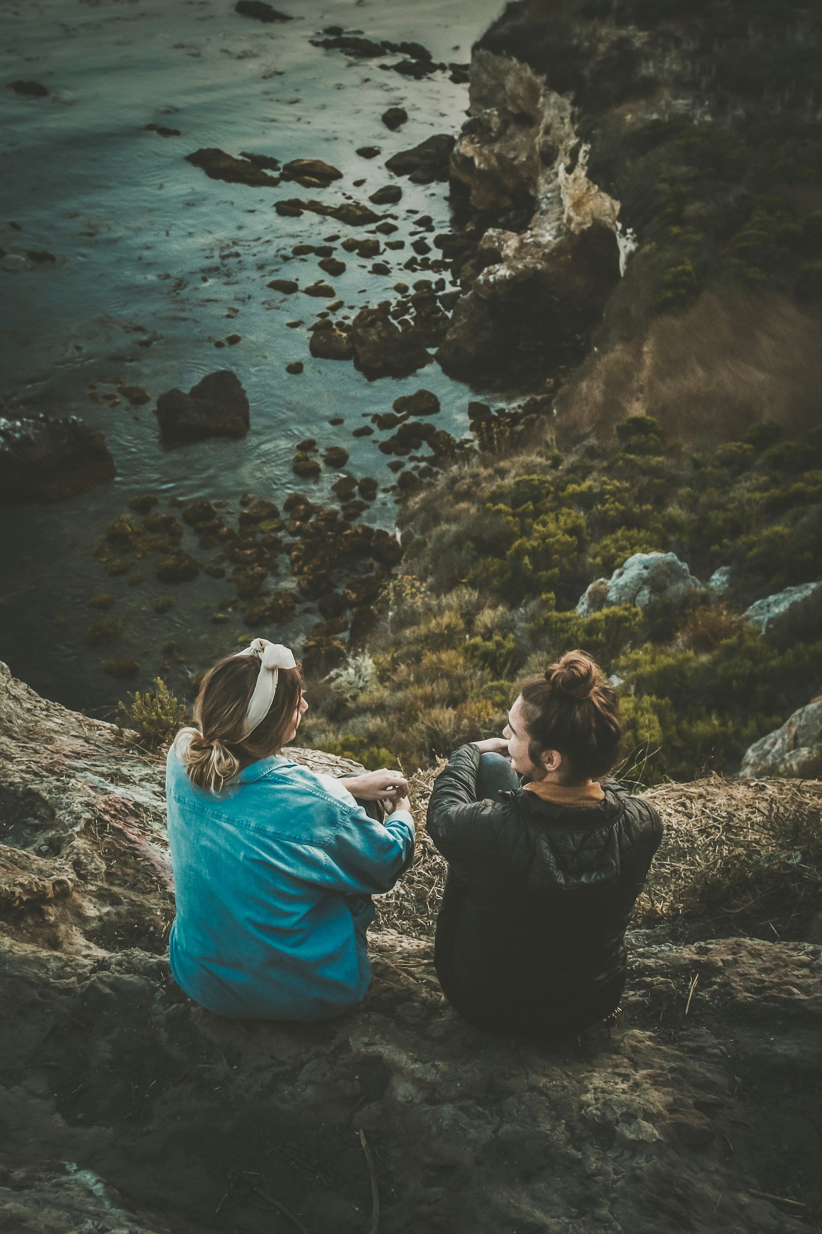 Two women sitting on rocks near a cliff and ocean, engaged in conversation, with rocky shoreline and cliffs in the background.
