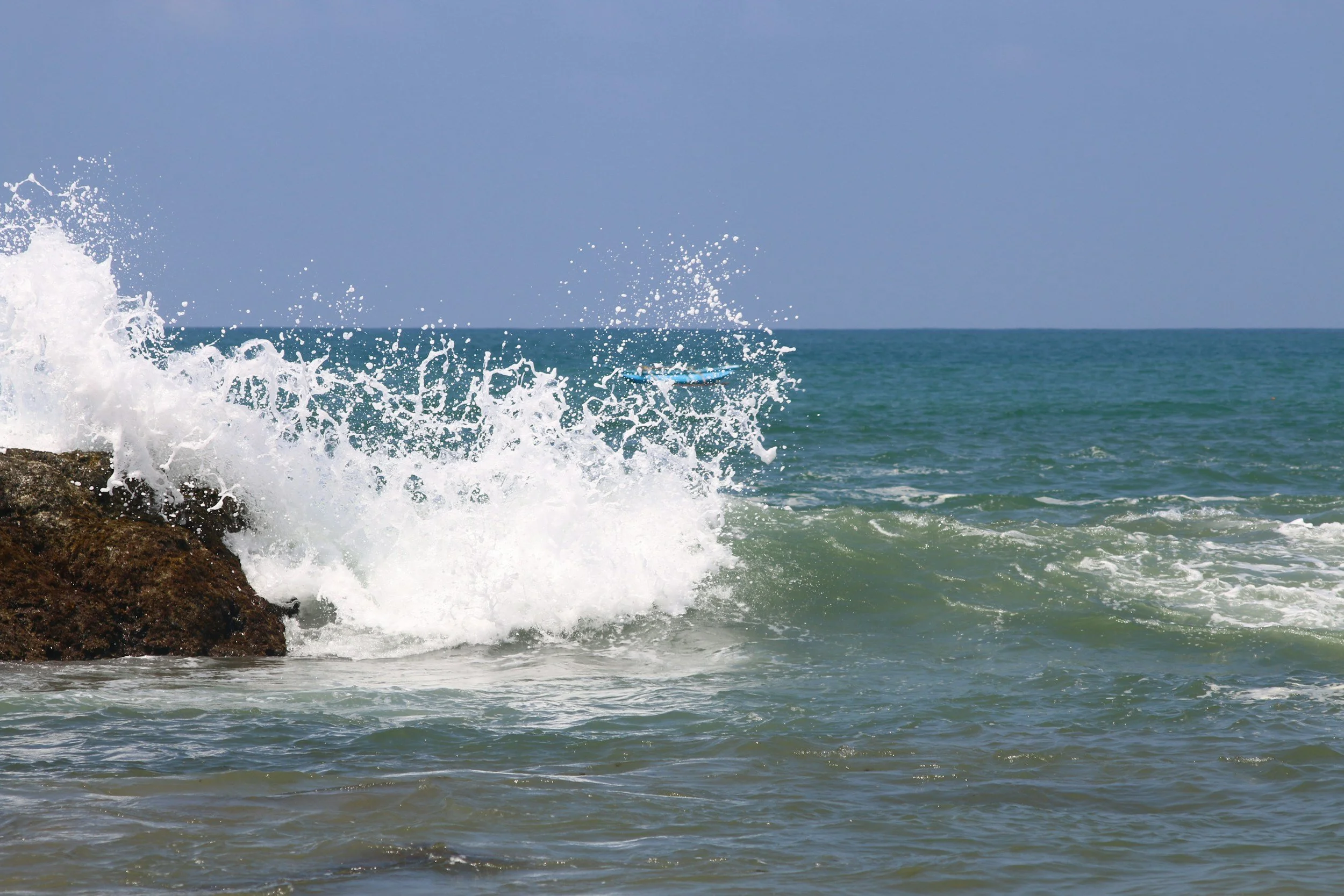Wave crashing against rocks on the shoreline with ocean and sky in the background.