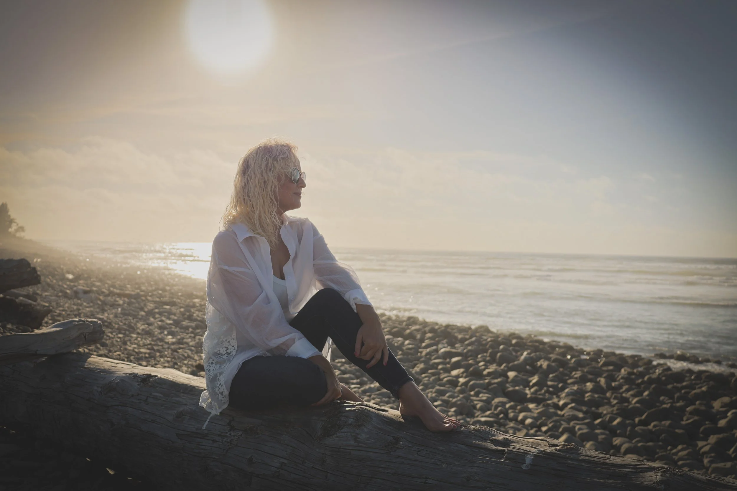 Founder of The Deep Current Collective sitting on driftwood overlooking the Oregon Coast at sunset.