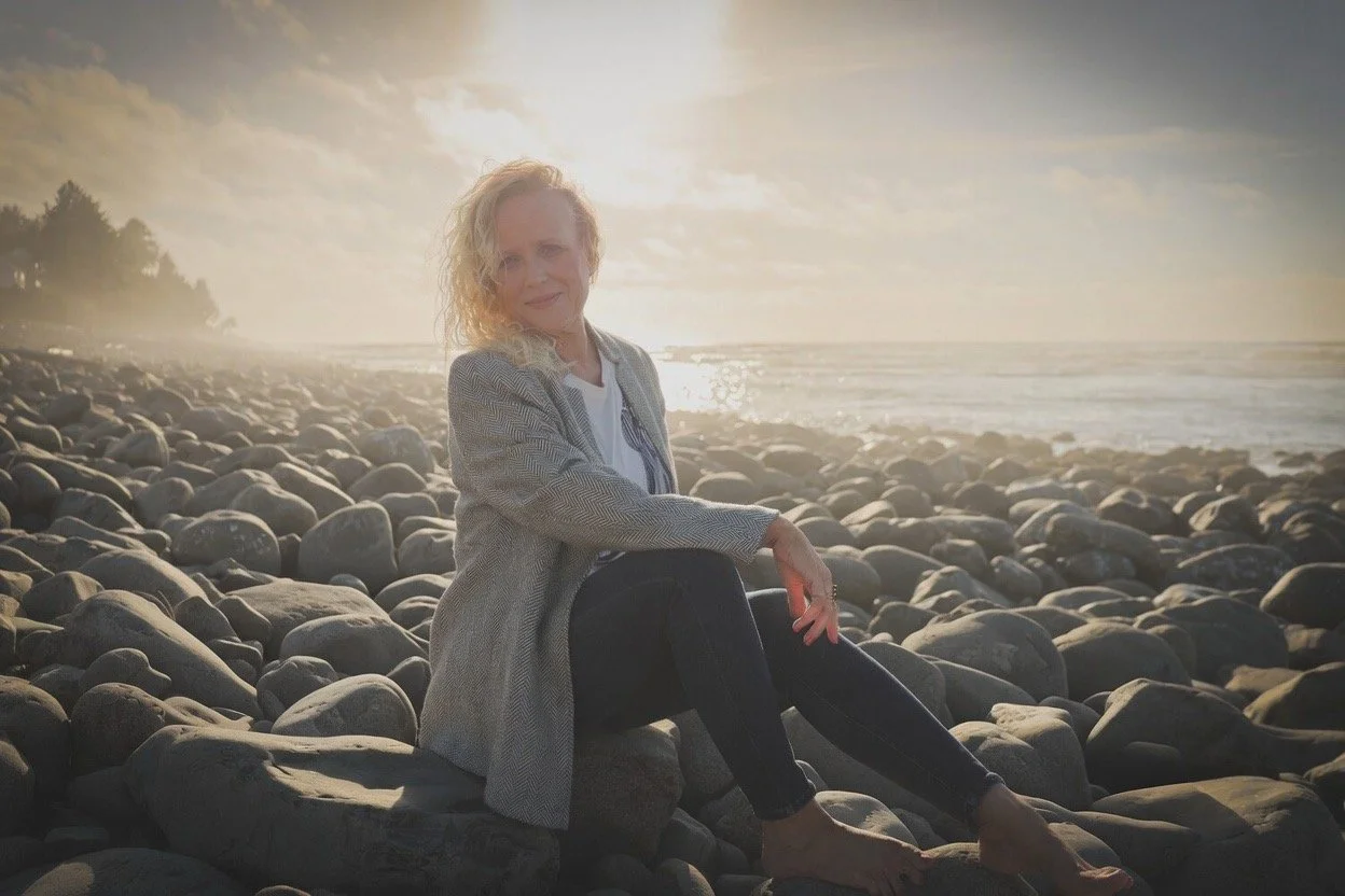 Founder of The Deep Current Collective sitting on rocky Oregon coastline at sunset.