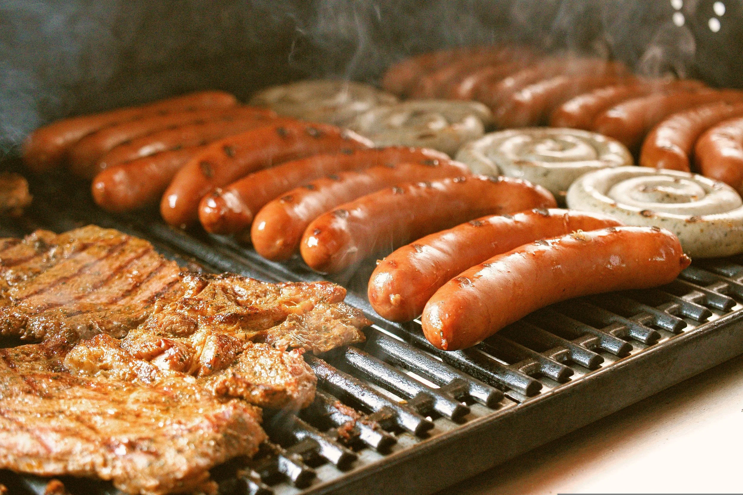 Sausages and a grilled meat patty cooking on a barbecue grill with smoke.
