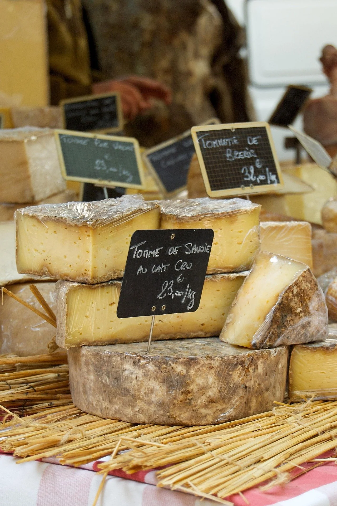 Various wheels of cheese on display at a market, with small chalkboard signs indicating their names and prices.