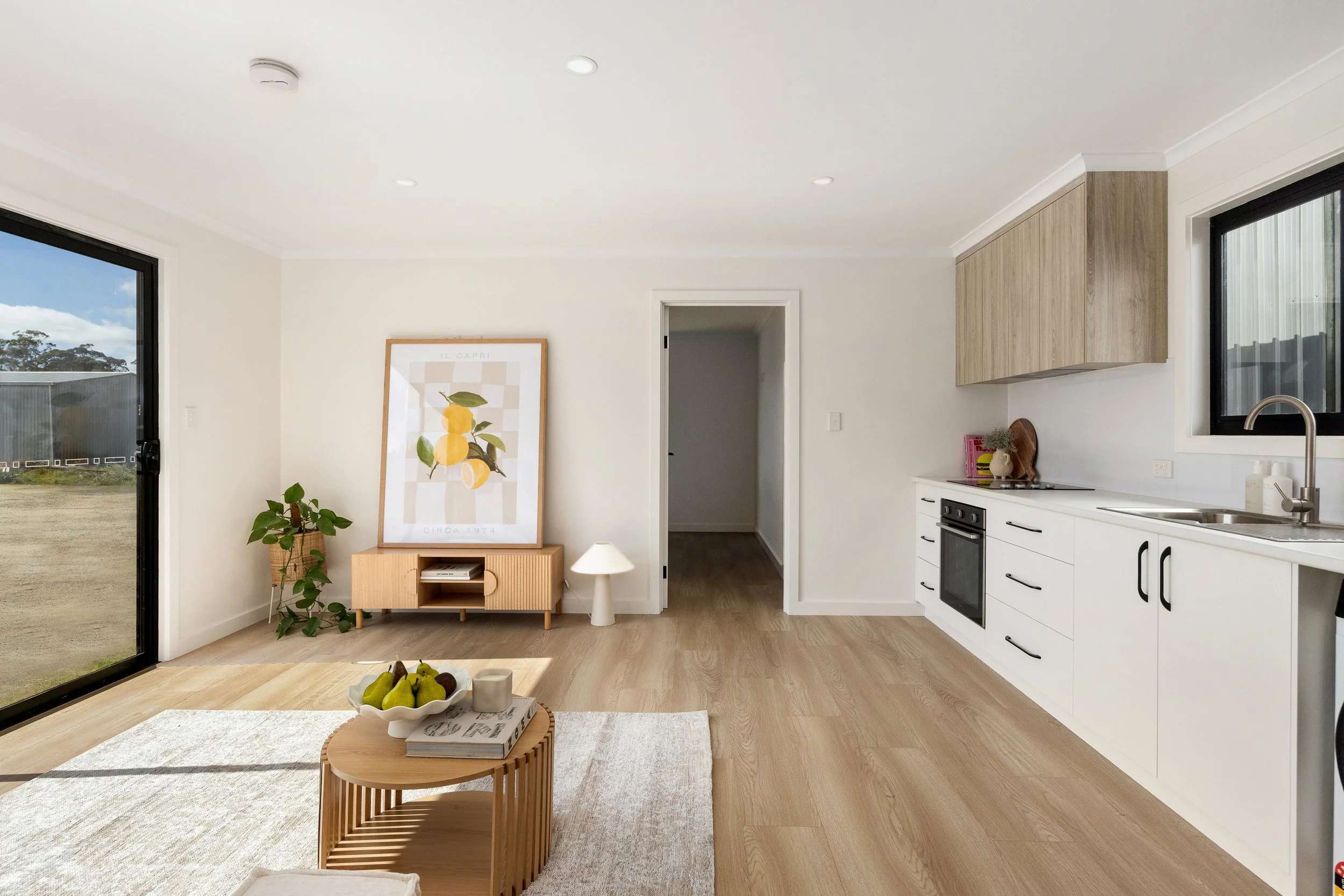 A bright, modern living room with a kitchen area. Features include large windows with black frames, a white ceiling with recessed lighting, a small wooden sideboard with a framed lemon print and a potted plant, a white floor lamp, a small round wooden coffee table with a bowl of pears and books, and a beige area rug. The kitchen has white cabinets, black handles, a black stove, a stainless steel sink, and a window above the sink.