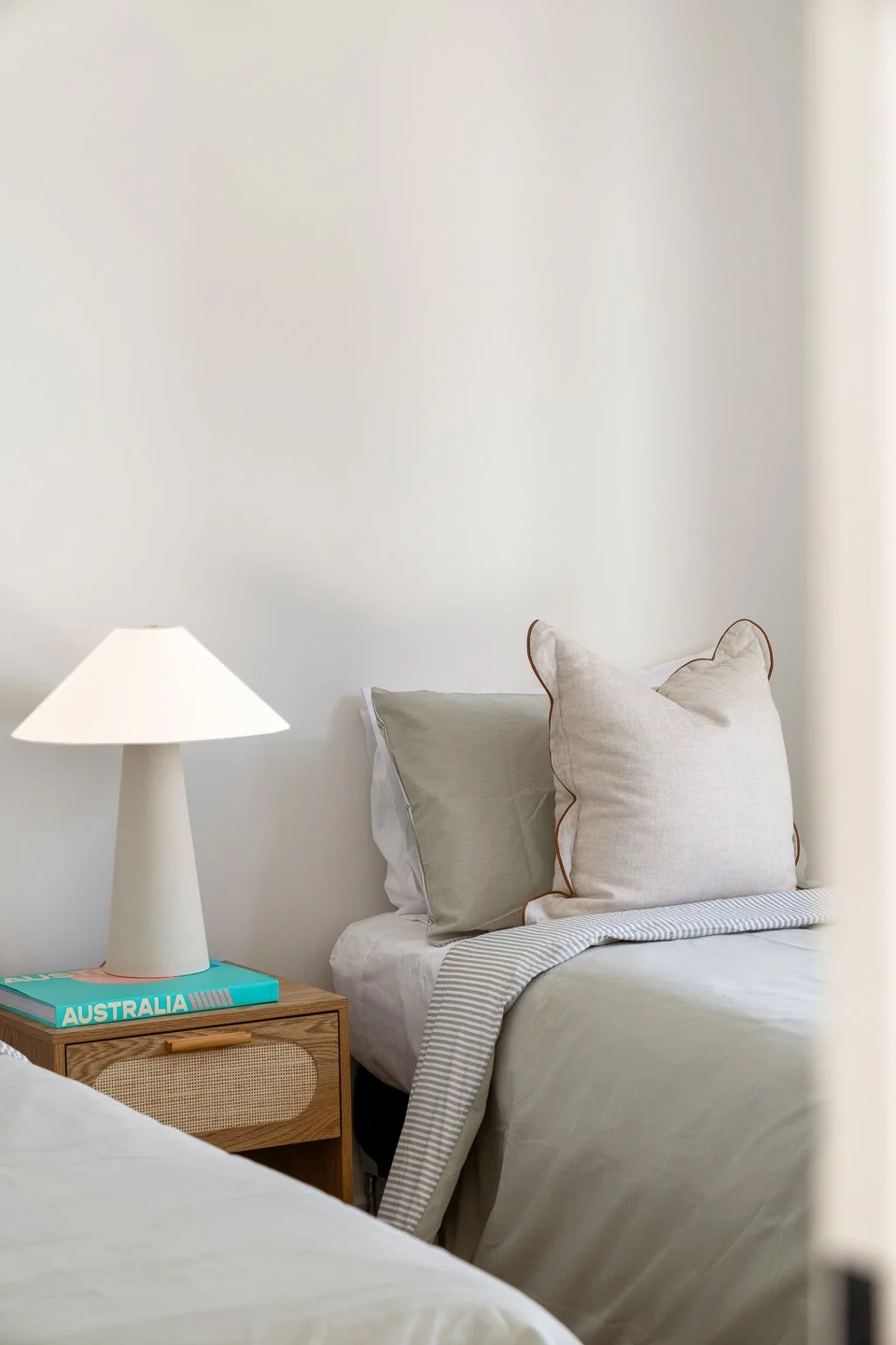 A cozy bedroom with neutral tones, a bedside table holding a modern white lamp and a teal book titled 'Australia,' and a bed with beige and gray pillows and a striped blanket.