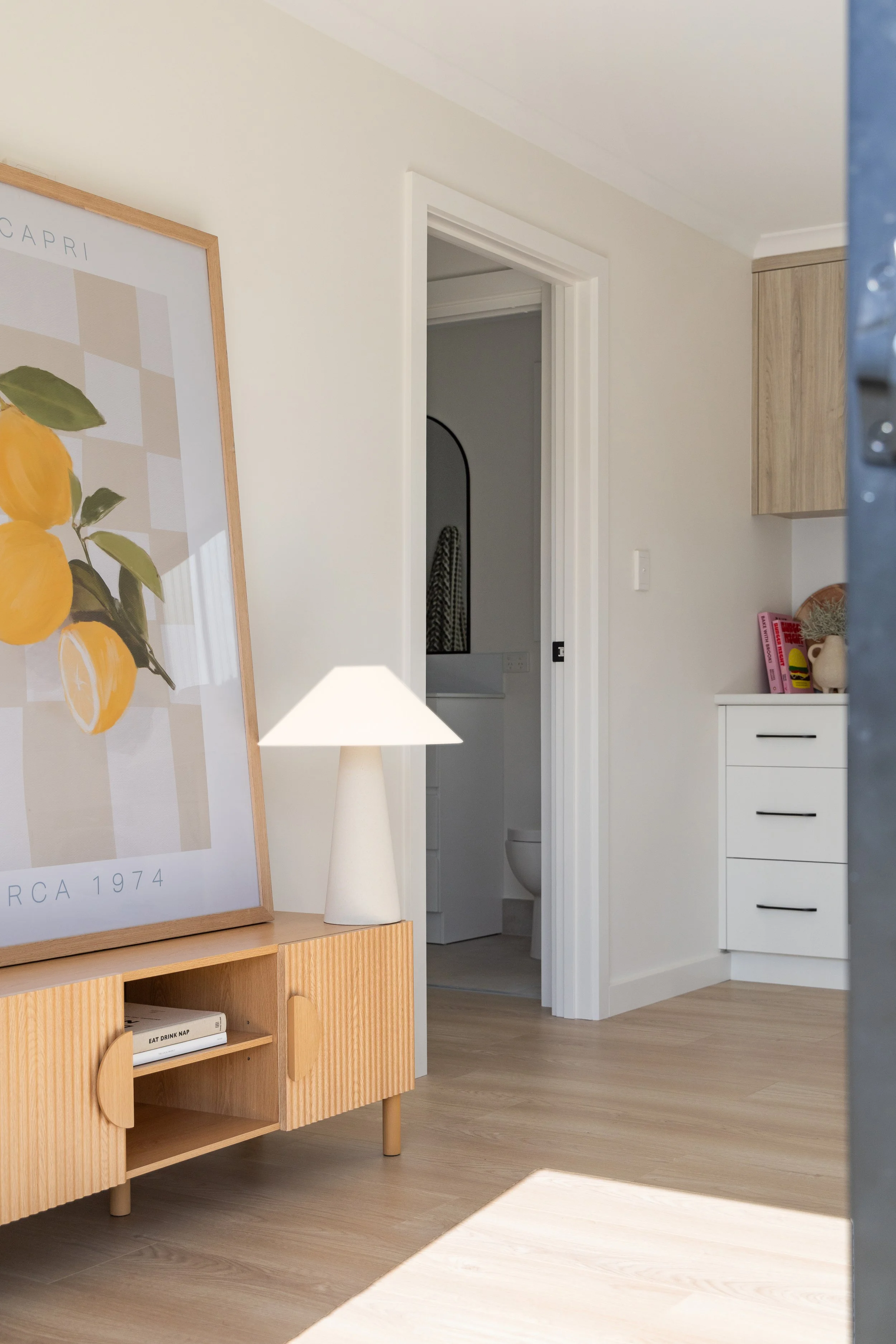 A view of a corner in a modern home interior, showing a white door open to a bathroom with a toilet, a wall-mounted mirror, and a tiled wall. Part of a wooden cabinet and a white dresser with books and decorative items are visible. There is a framed art print of lemons and a white lamp on a wooden surface.