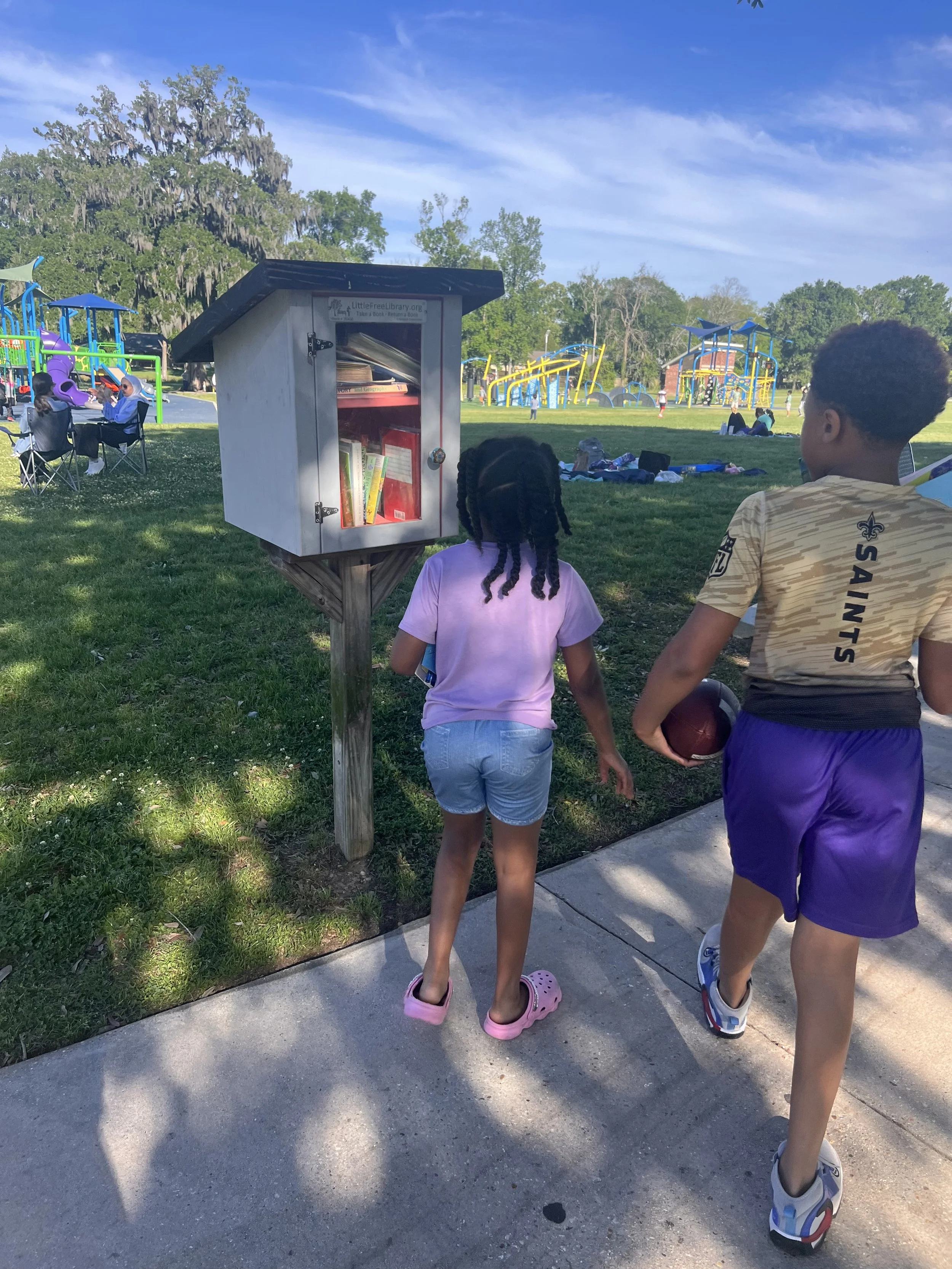 Children walking on sidewalk near a park with a book exchange box and playground in the background.