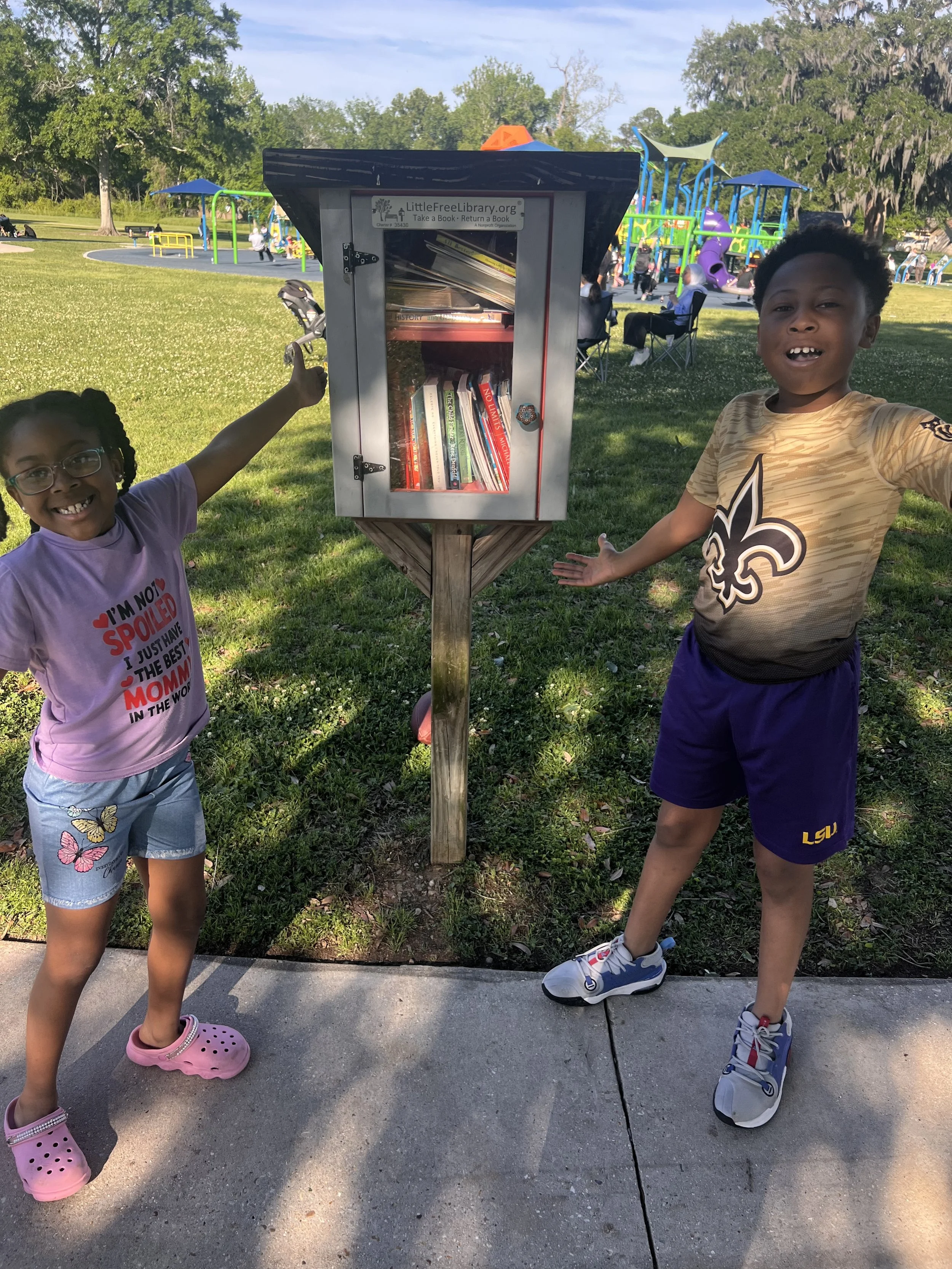 Two children, a girl and a boy, stand on a sidewalk next to a small outdoor library box filled with books. The girl has glasses and is smiling, wearing a pink shirt and butterfly-patterned shorts, with pink Crocs. The boy is wearing a sports jersey a