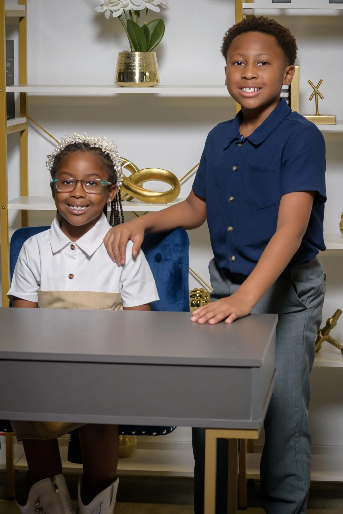 Two children, a girl seated and a boy standing, smiling at the camera in a modern room with white walls, gold accent decorations, and a bookshelf with plants.