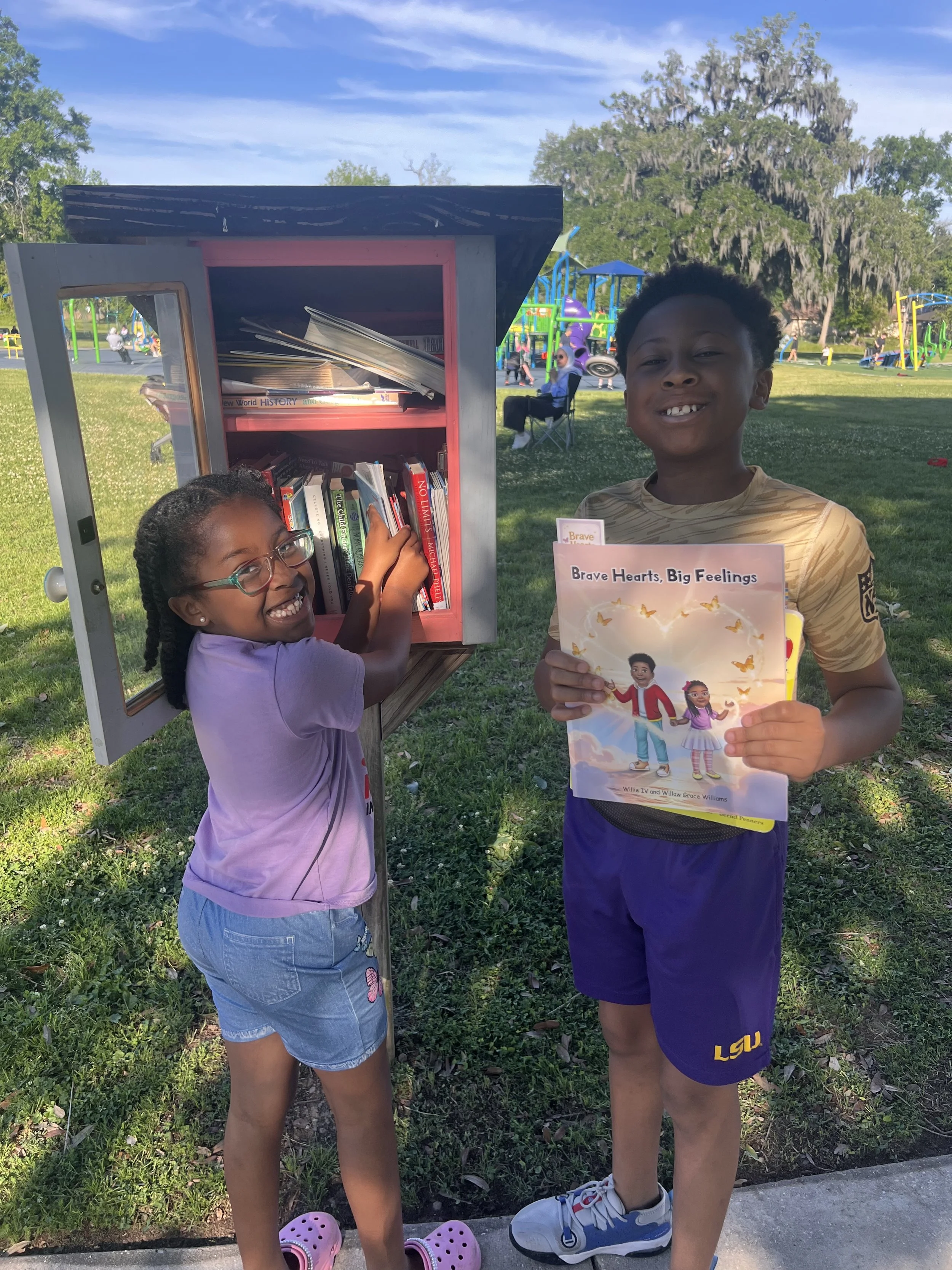Two children, a girl and a boy, are at a park in front of a small gray and pink library box with books inside. The girl is wearing glasses, a purple shirt, and denim shorts, and is reaching into the box. The boy is holding a book titled 'Brave Hearts