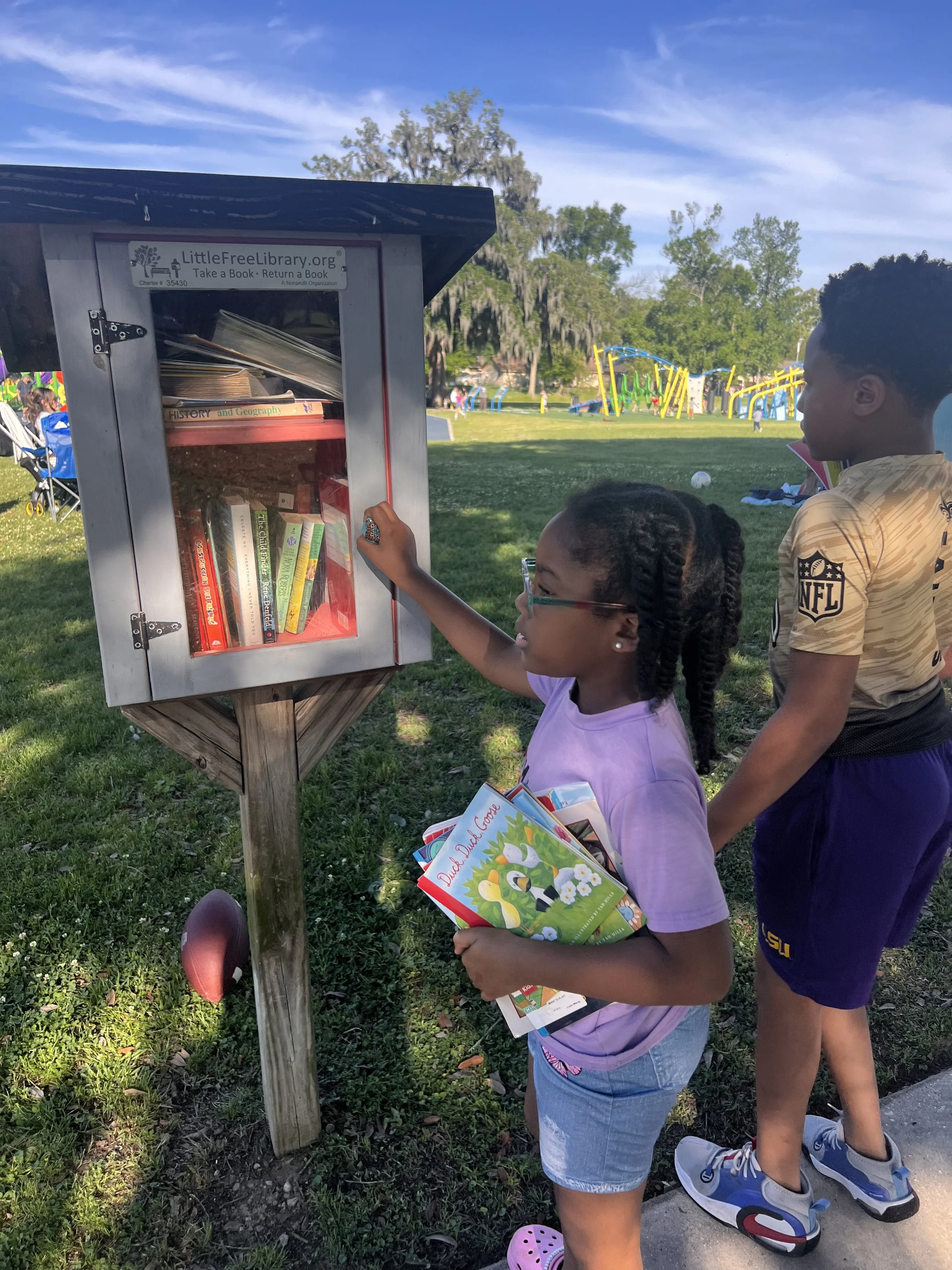 Two children, a girl and a boy, stand in front of a Little Free Library outdoors in a park. The girl is holding children's books, and the boy is looking into the library box on a post, which contains various books and magazines. The park has grass, t