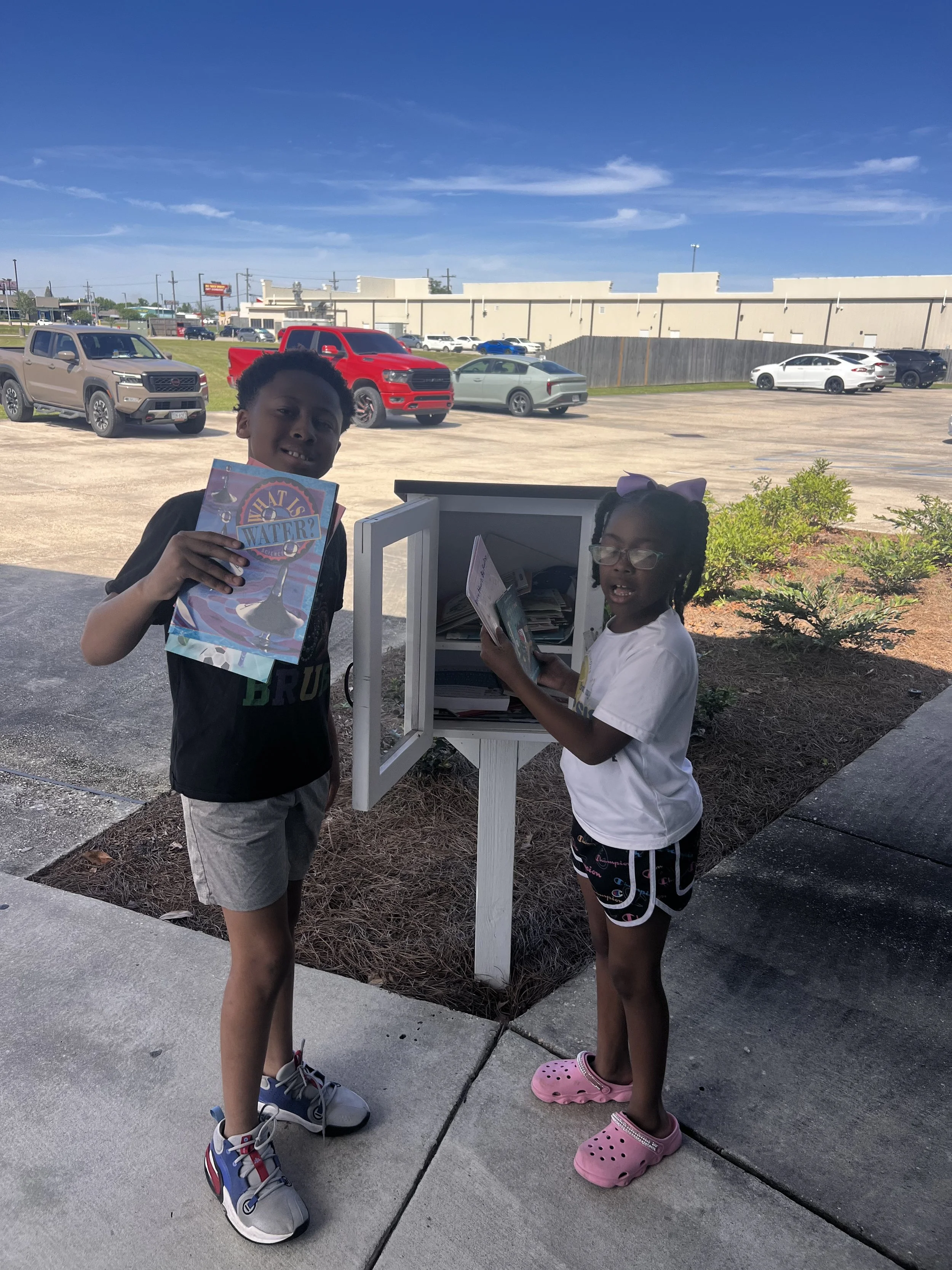 Two children standing outside near a mailbox, reading books with cars and a building in the background under a blue sky.