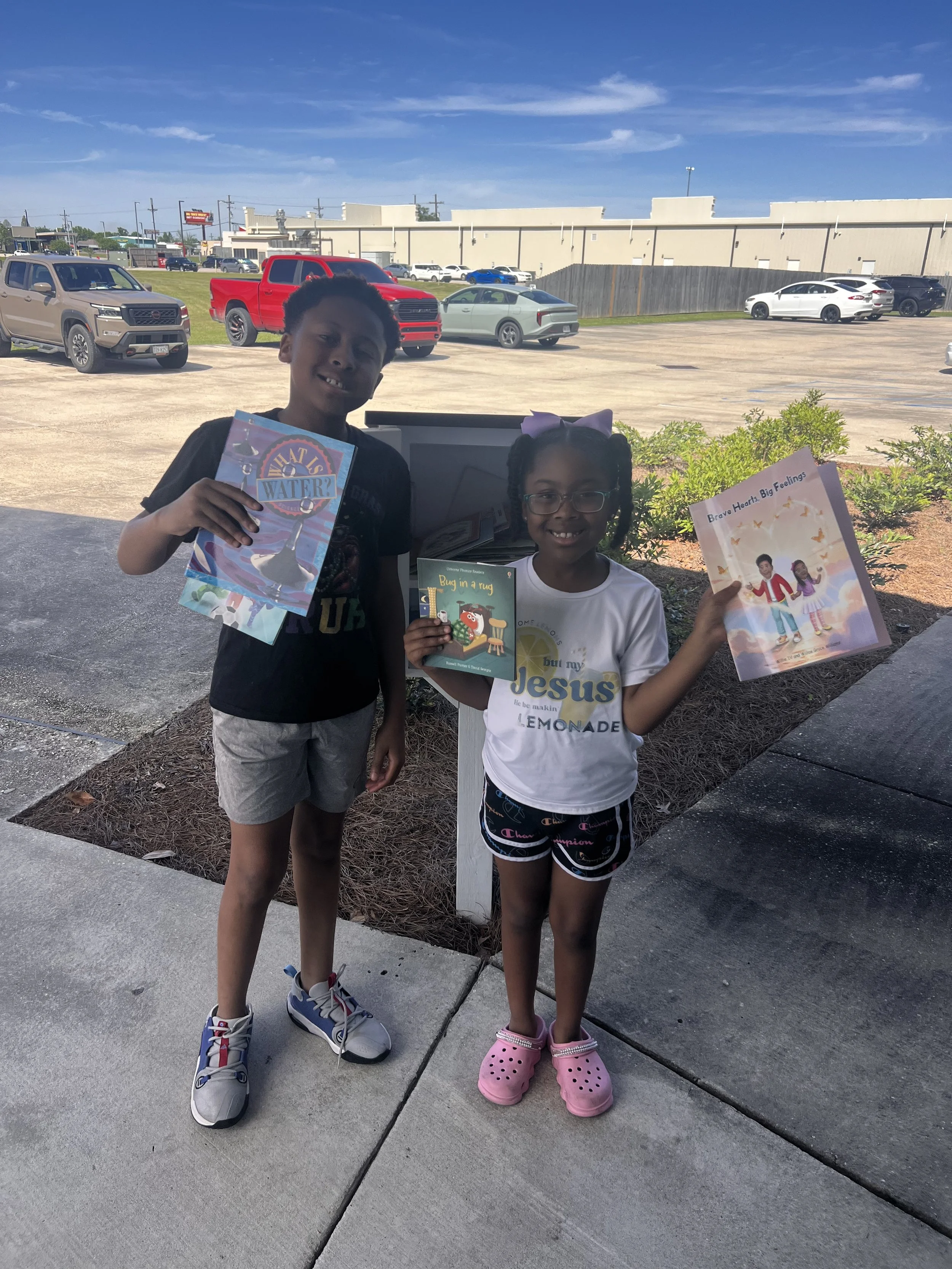 Two children, a boy and a girl, are standing outdoors in front of a parking lot with several cars. The boy is holding a colorful book titled 'What is Water?' and is wearing a dark t-shirt, shorts, and sneakers. The girl is smiling, wearing glasses, a