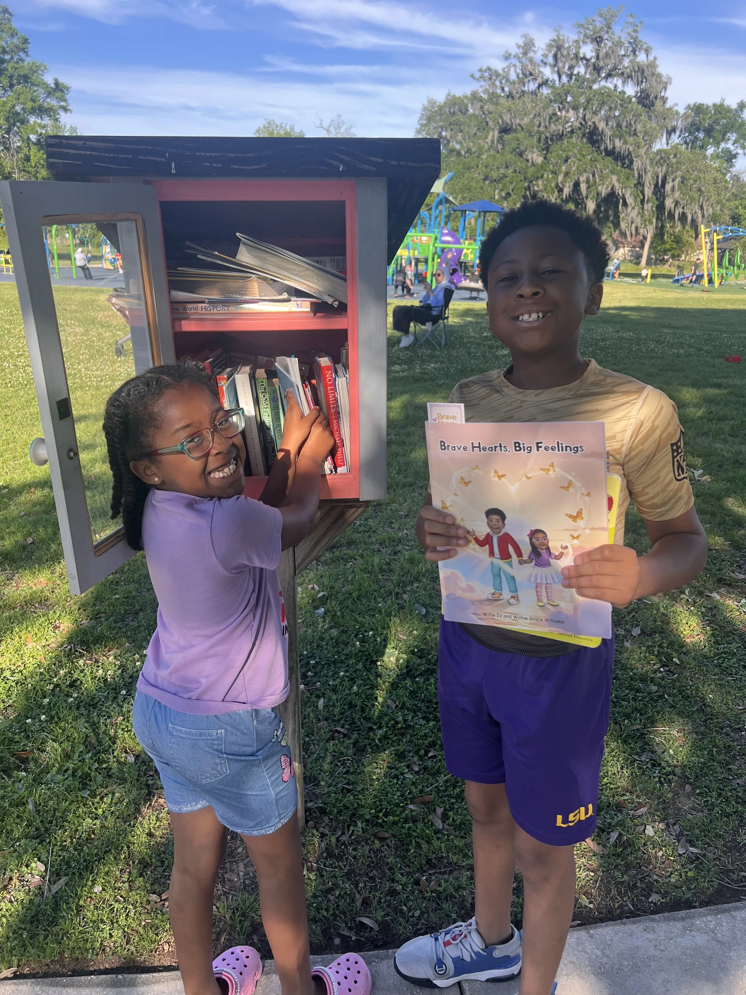 Two children, a girl and a boy, are smiling outdoors near a small bookshelf in a park. The girl is wearing a lilac shirt, denim shorts, and pink Crocs, reaching into the bookshelf. The boy, wearing a LSU sports shirt and purple shorts, is holding a book titled "Brave Hearts, Big Feelings". The park has playground equipment and large trees in the background, with other people visible.