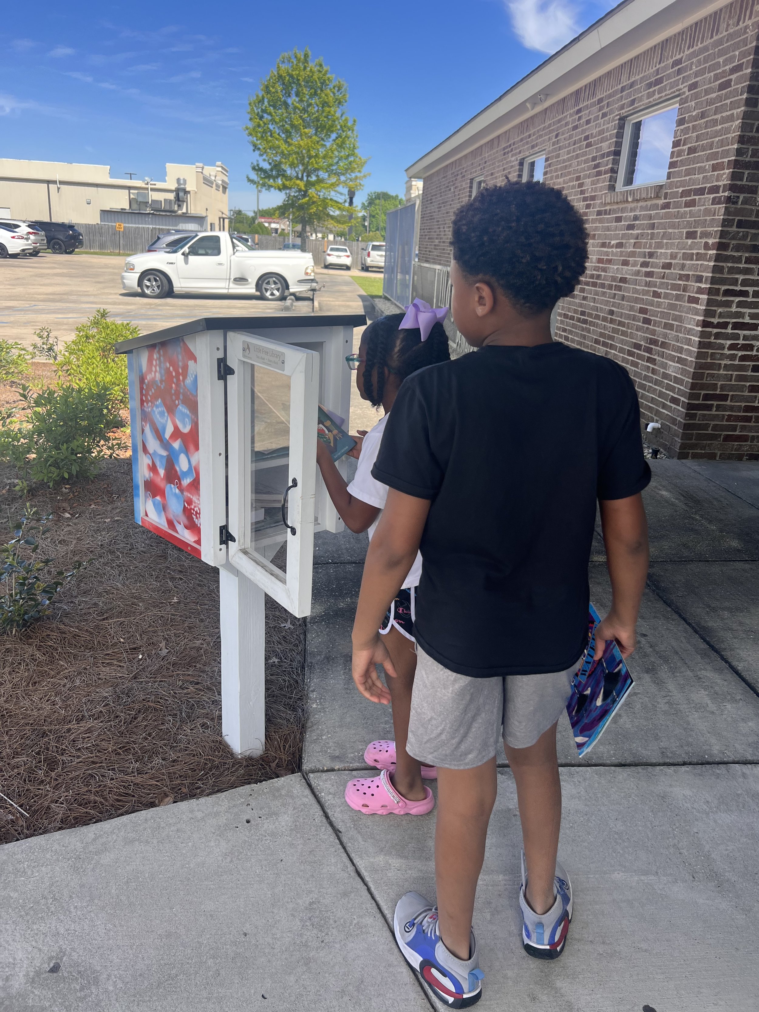 Two children, a girl and a boy, standing in line at a transparent kiosk outside near a brick building, with parked cars and a blue sky in the background.