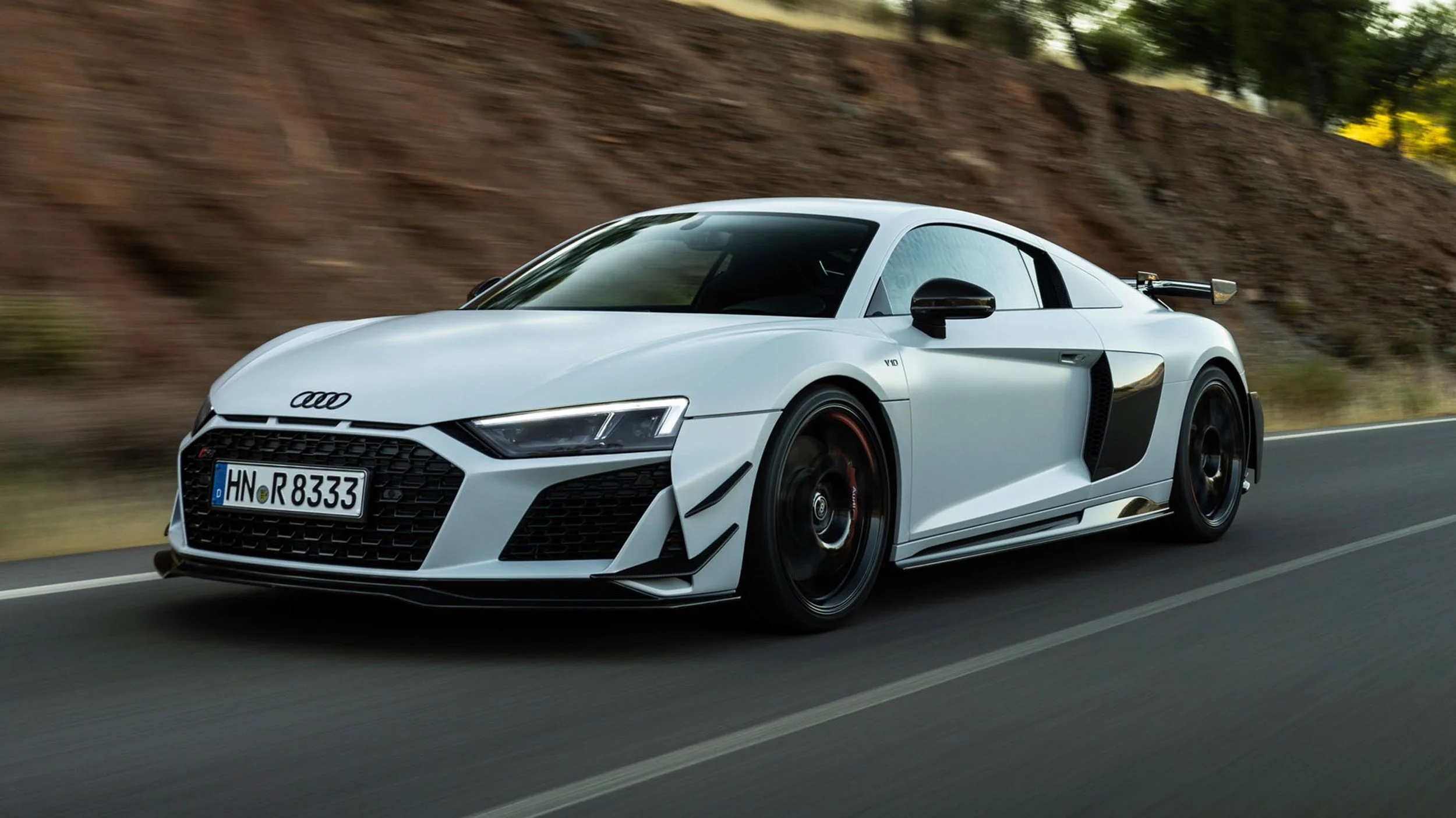 White Audi sports car driving on a highway with blurred background of trees and rocks.