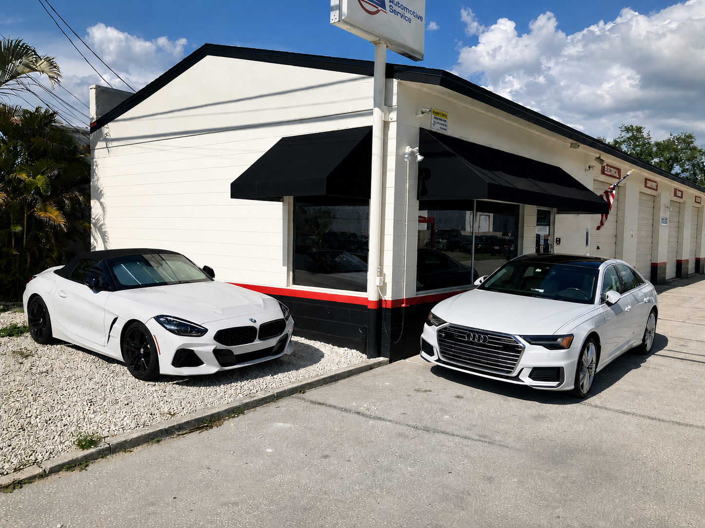 White BMW convertible and white Audi sedan parked outside an automotive service shop with black awnings and a sign.