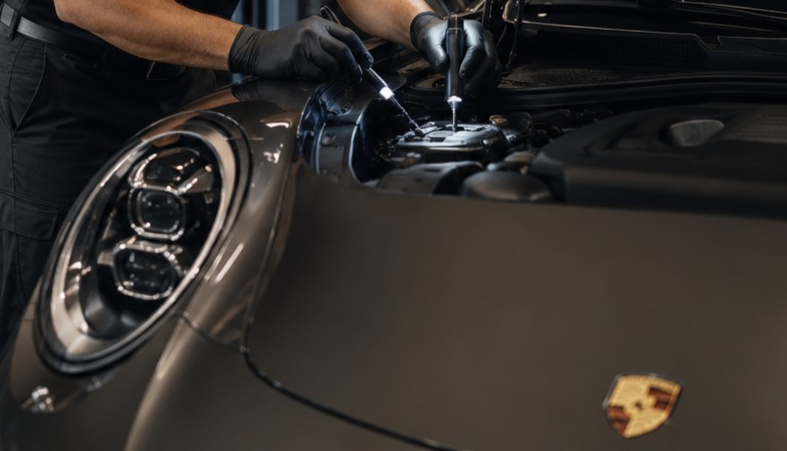 A mechanic wearing black gloves performing maintenance or repairs under the hood of a black Porsche car.