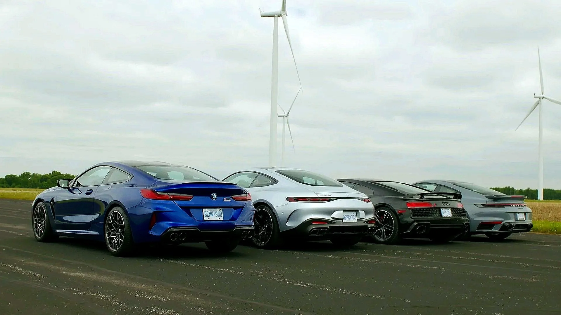 Four sports cars parked on an open road with wind turbines in the background under a cloudy sky.