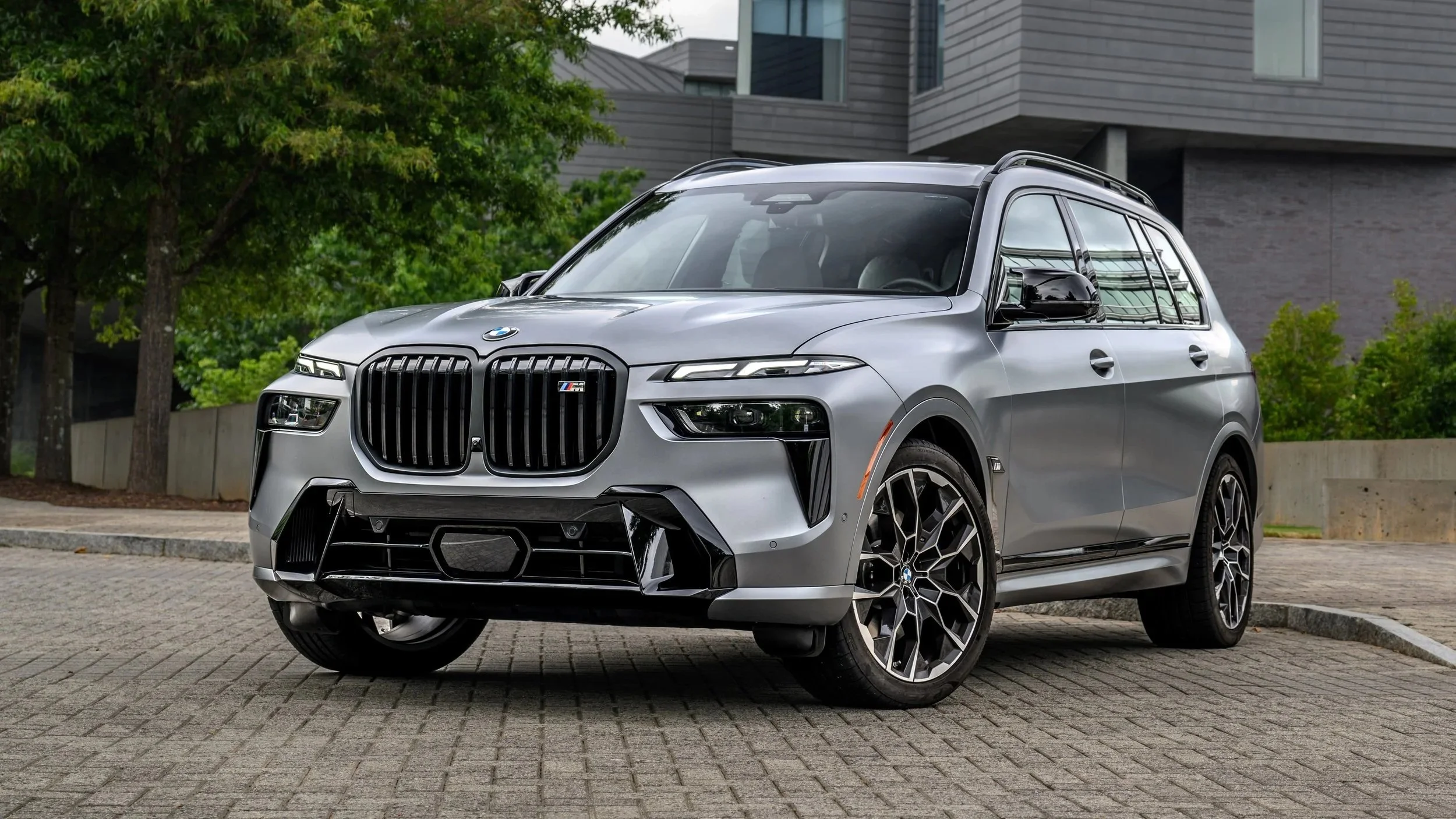 A silver BMW SUV parked on a brick lot with trees and modern buildings in the background.