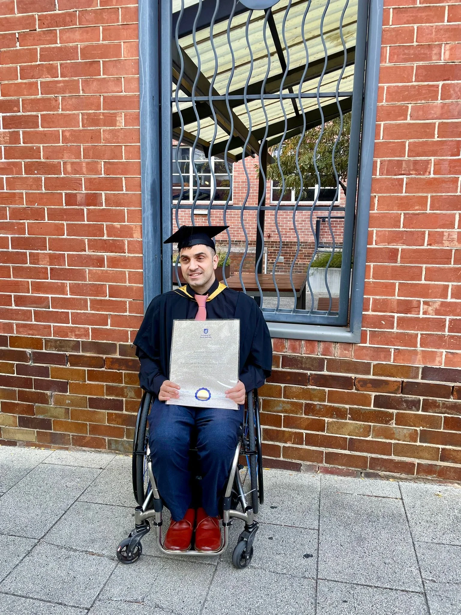 A man in a wheelchair wearing academic regalia holding a diploma standing in front of a brick building with a metal fence and stairs in the background.