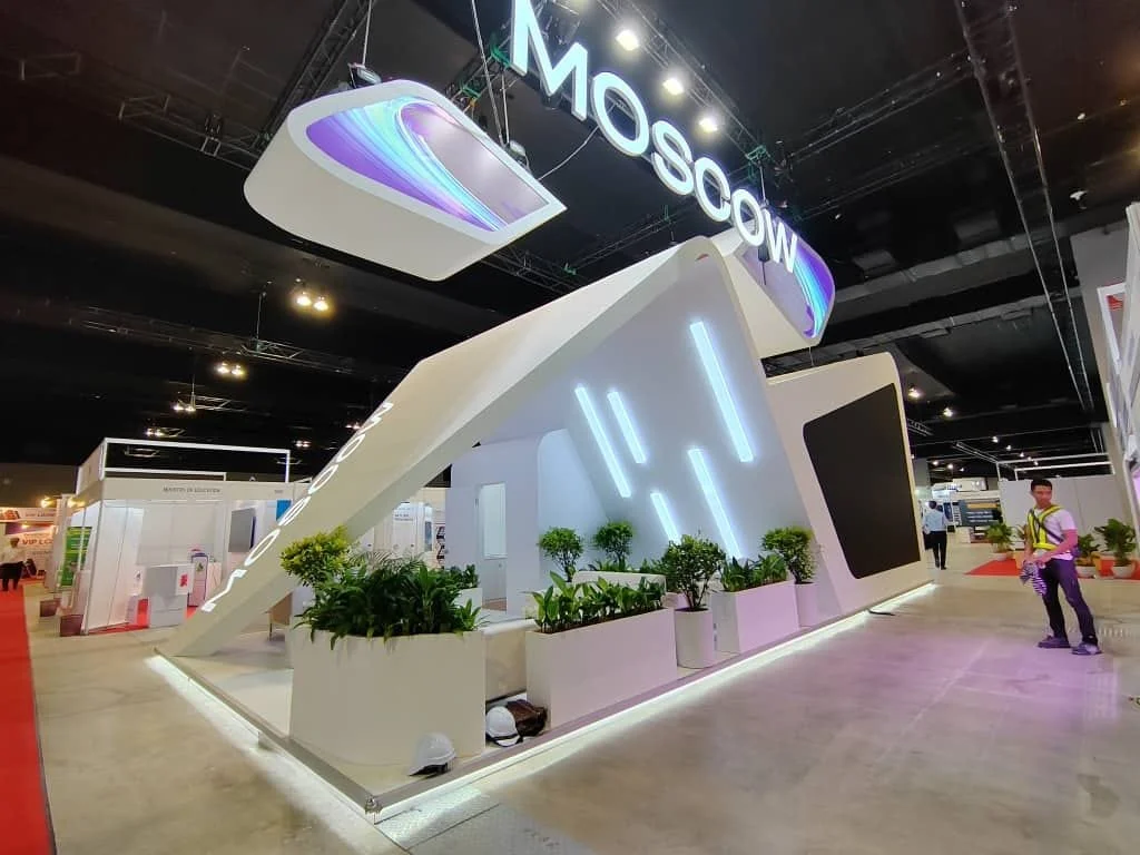 Exhibition booth labeled 'Moscow' with modern white structure, illuminated vertical lines, and potted plants, at an indoor convention with people walking around.