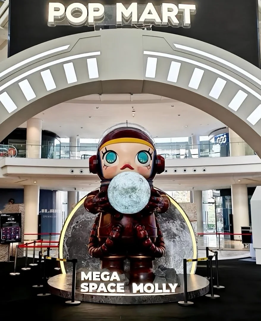 A large cartoonish sculpture of a girl with big blue eyes, wearing a space helmet, holding the moon, with the words 'MEGA SPACE MOLLY' below at an indoor shopping mall or entertainment center.