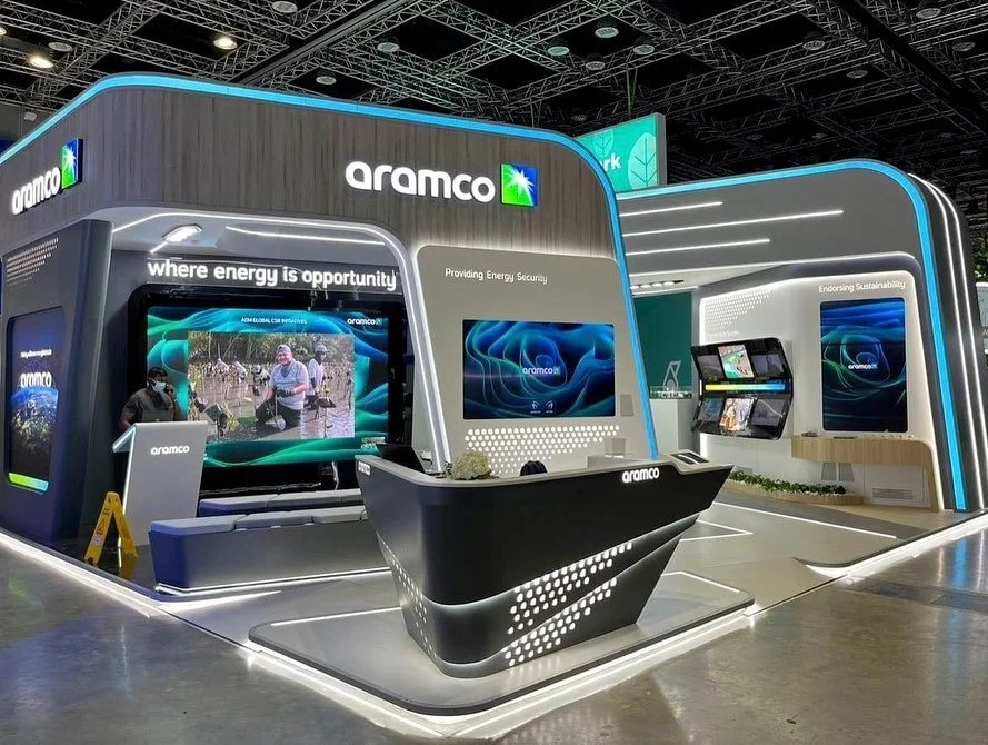 Exhibition booth for Aramco featuring large digital screens, a reception desk, and branding with blue and green accents at a convention center.