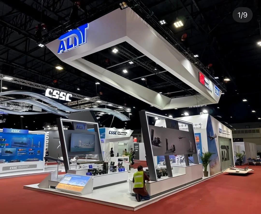 Trade show booth for ADI and CSSC featuring displays, screens, and model ships, with a red carpeted floor and black ceiling trusses.