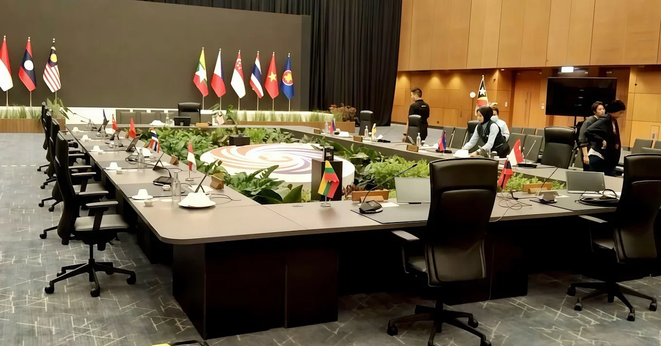 Conference room with U-shaped table setup, surrounded by black chairs, with flags of various countries displayed behind the table, and people preparing the room.