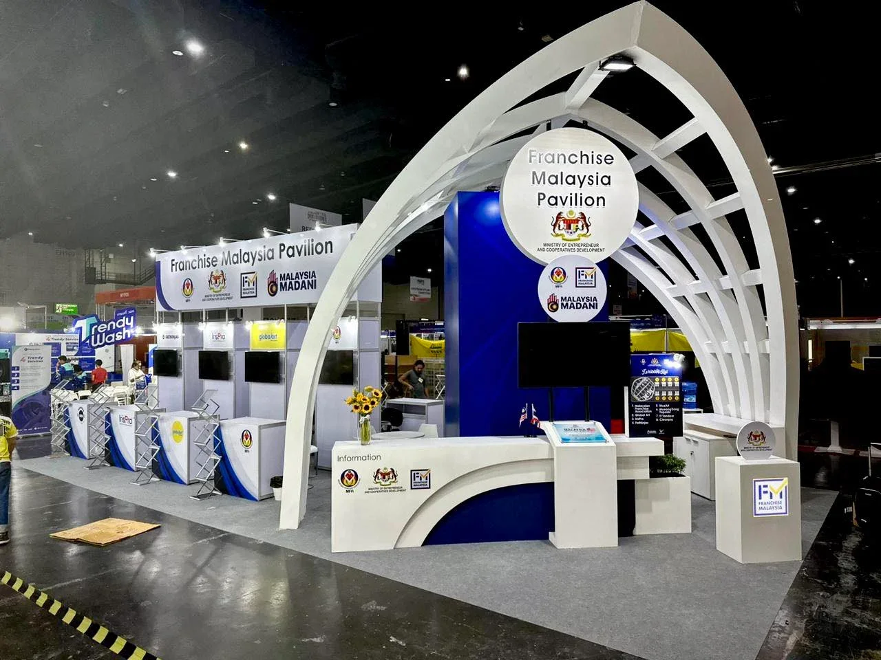 Exhibition booth for Franchise Malaysia Pavilion with white and blue design, featuring signage, display screens, and a reception counter, with a sign indicating it is part of the Ministry of Entrepreneur and Cooperative Development, Malaysia, at a trade event.