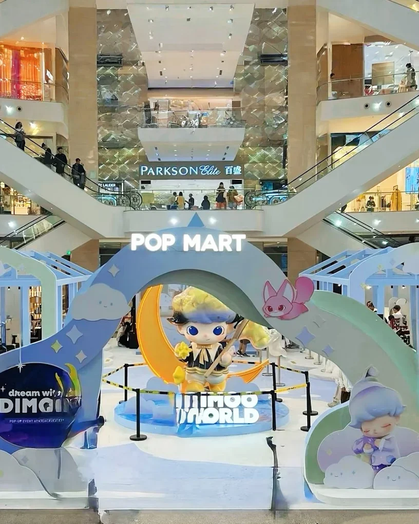 A Toyoo World display in a shopping mall with a cartoon character holding a fishing rod, surrounded by clouds, stars, and a pink bunny, with a sign that reads 'POP MART' and 'dream with DIMG' in the background.
