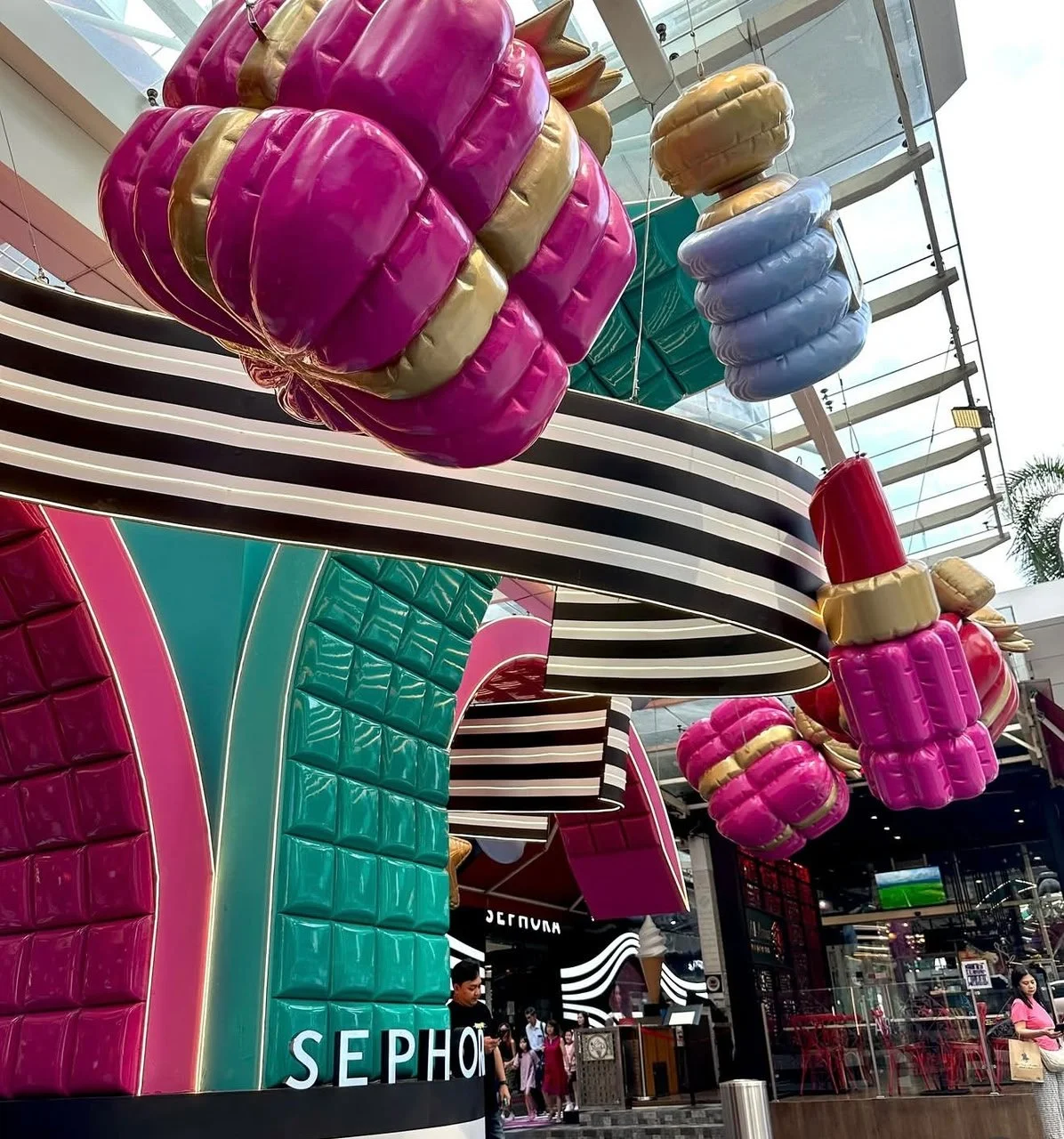 Colorful oversized sculptures resembling lipsticks and makeup items hanging inside a shopping mall with a Sephora store in the background.