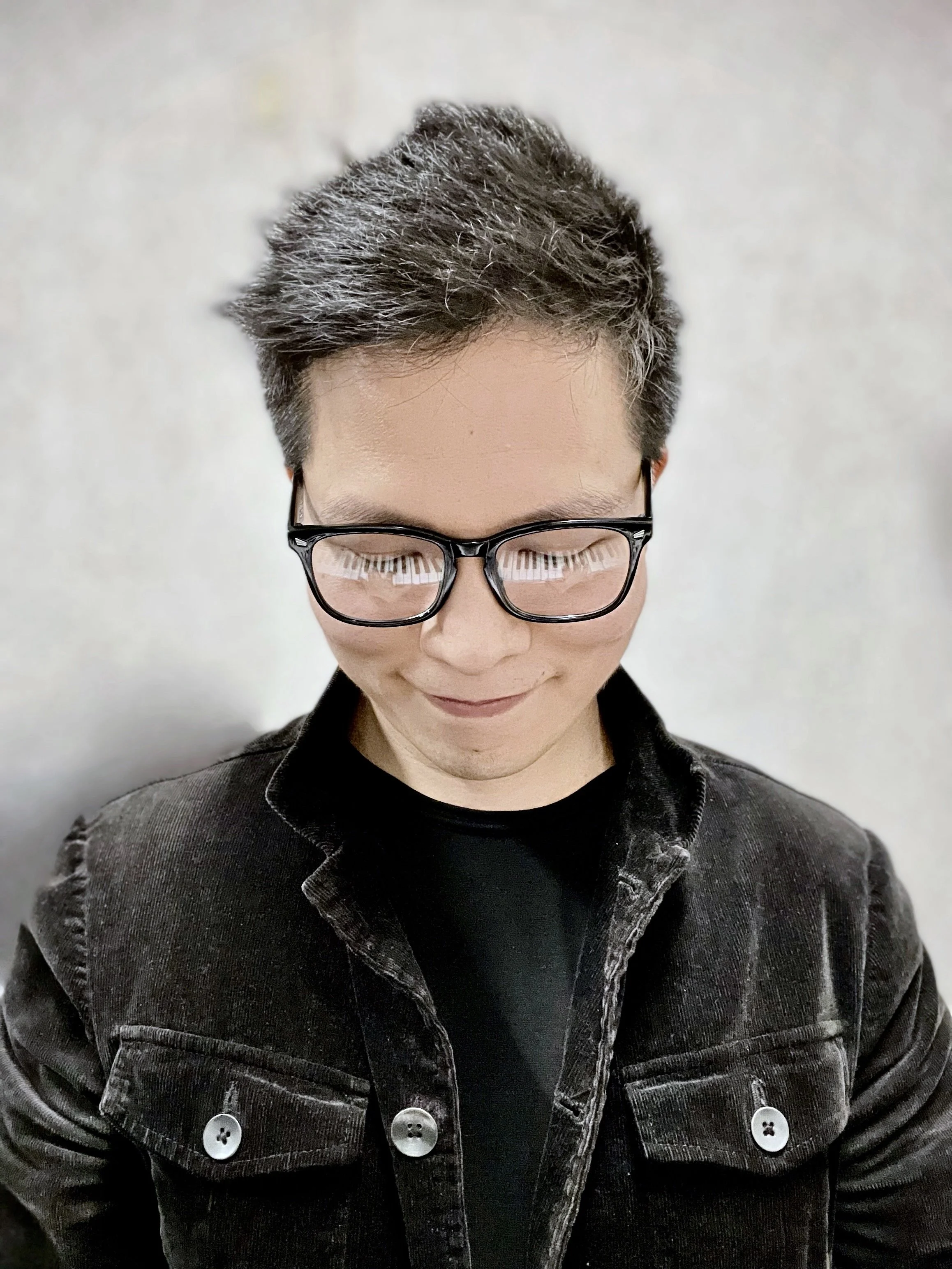 A person with short, gray and black hair, wearing glasses with a reflection of a keyboard on the lenses, and a black denim jacket with white buttons.