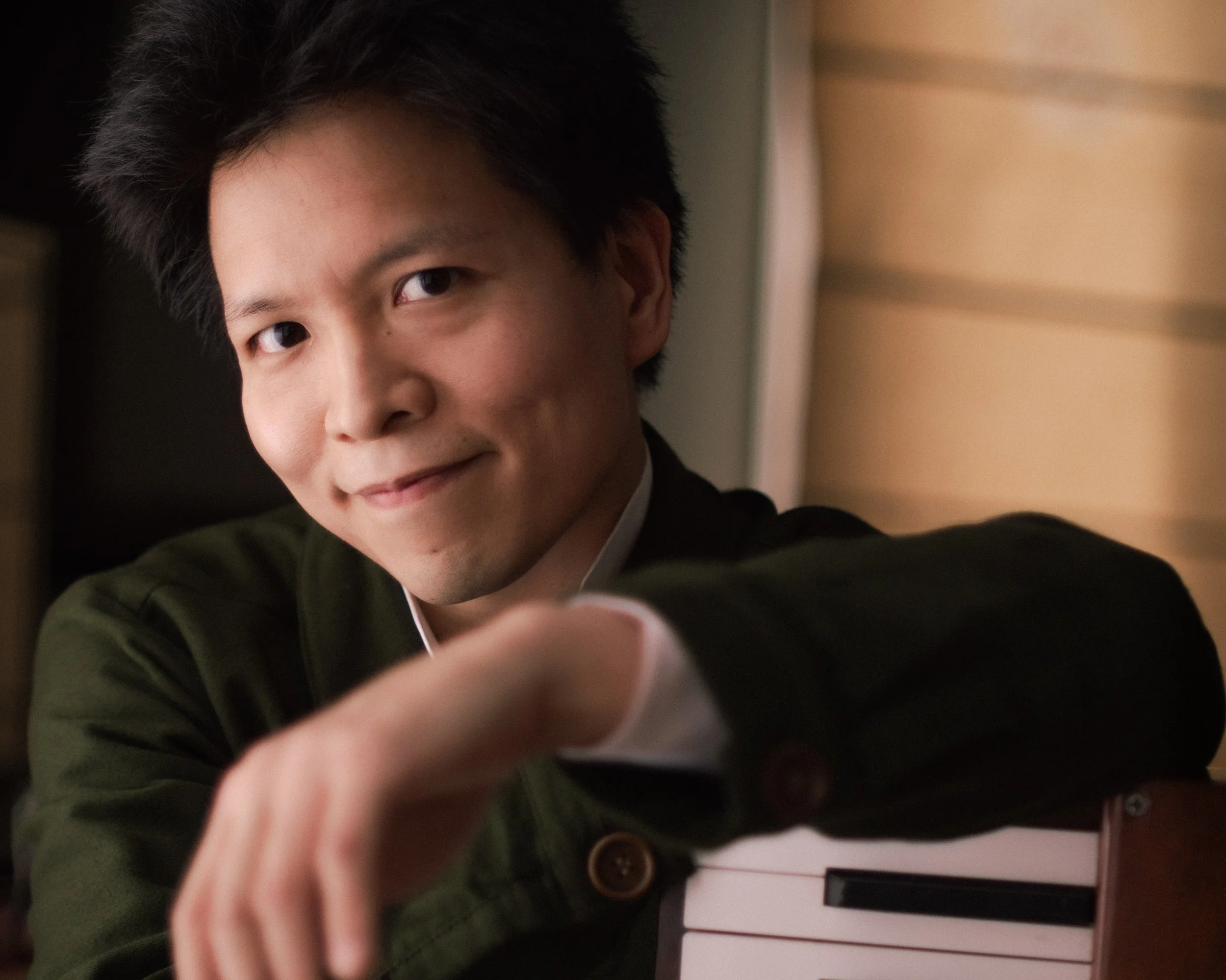 A close-up of a young man taking a selfie, smiling, with dark hair, wearing a green jacket, and a white shirt underneath. The background shows a blurred interior space with wooden panels and a white door.
