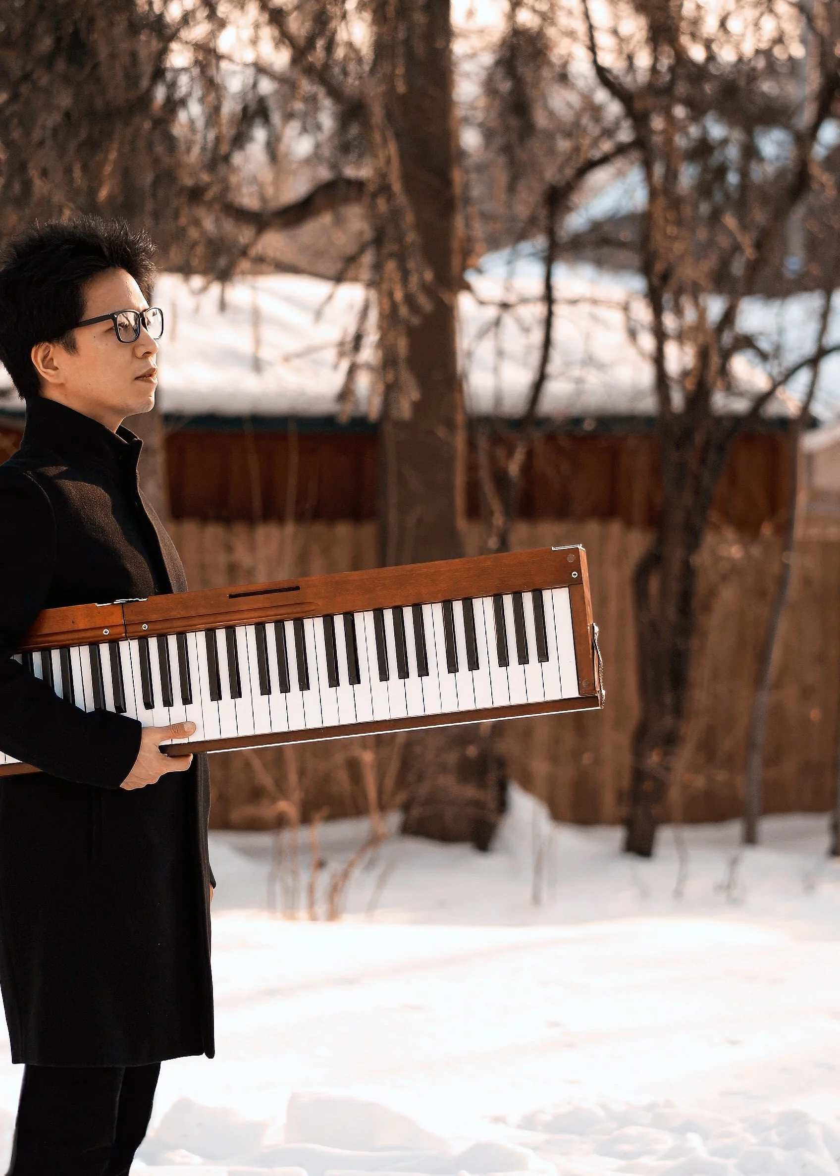 A person in a black coat and glasses stands outdoors in a snowy landscape, holding a wooden and black digital piano keyboard in front of them, with trees in the background.