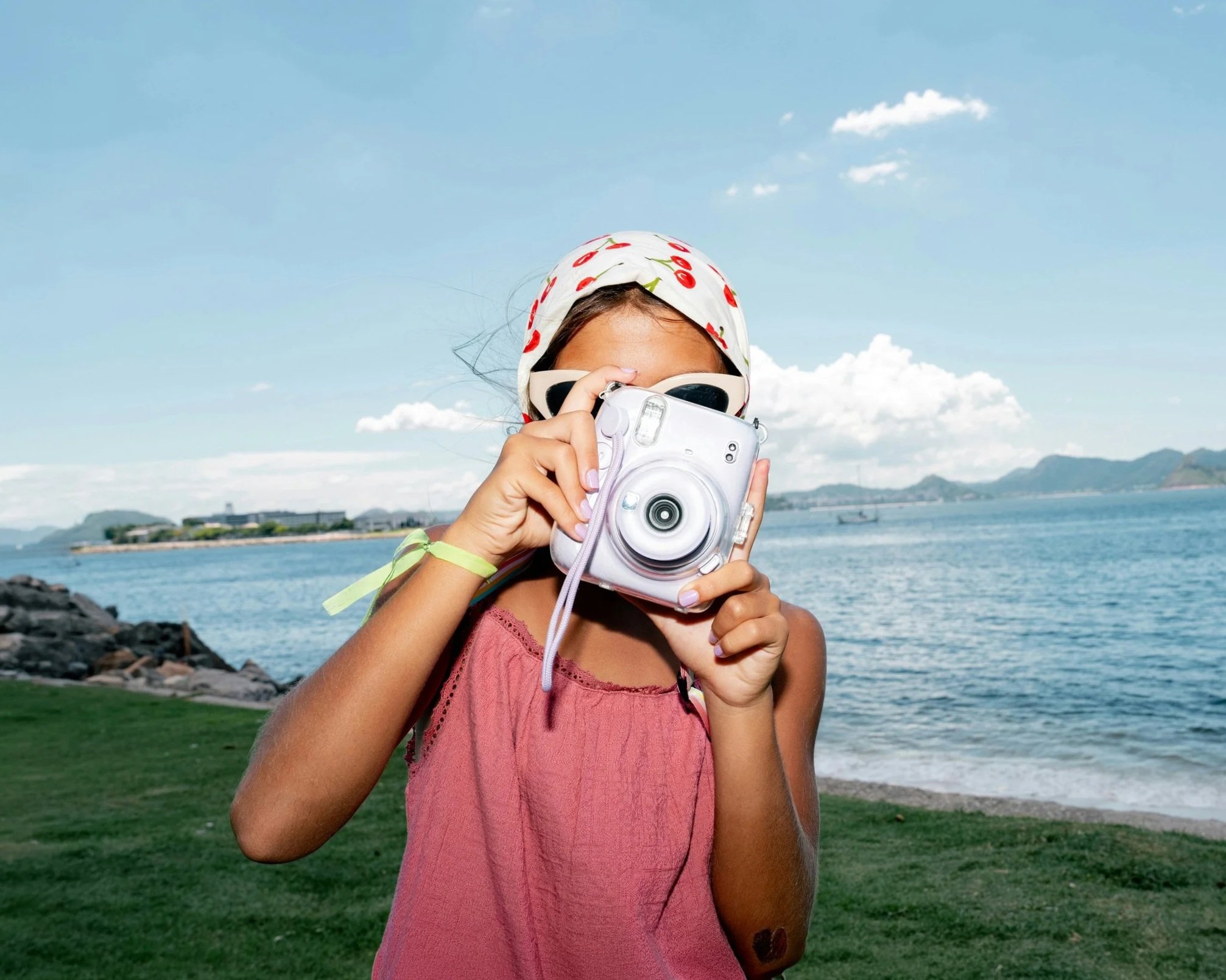A young girl wearing a pink sleeveless top and a white headscarf with red cherries is outdoors by a body of water, holding a camera and taking a picture.