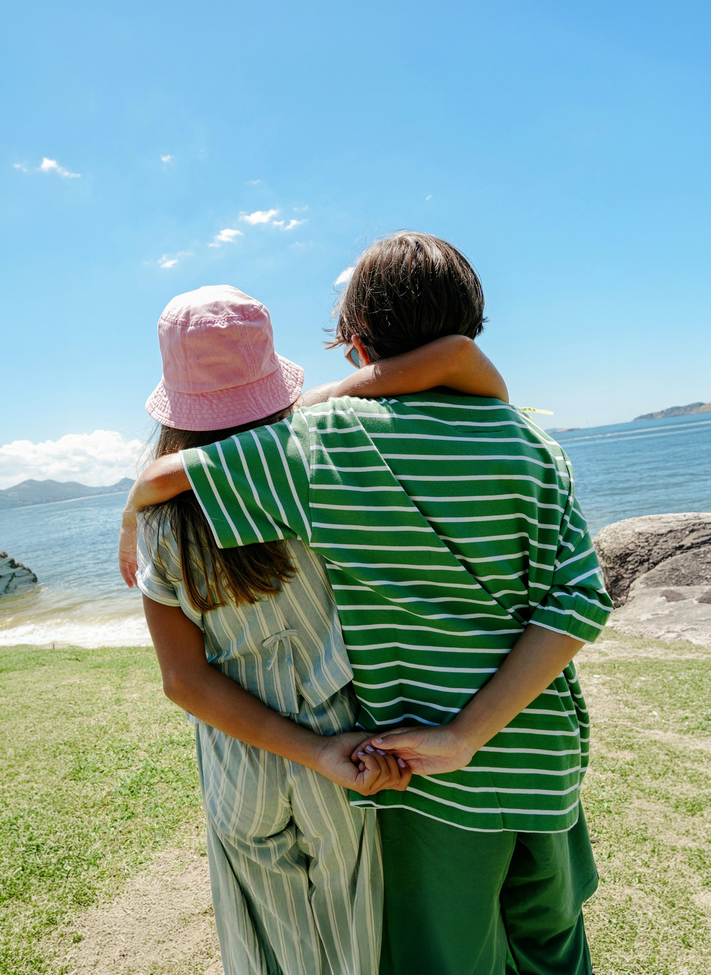 A couple stands close together on a grassy area near water, with the person on the right wearing a green and white striped shirt and the other person wearing a striped dress and pink hat, embracing each other while facing the ocean in the background.