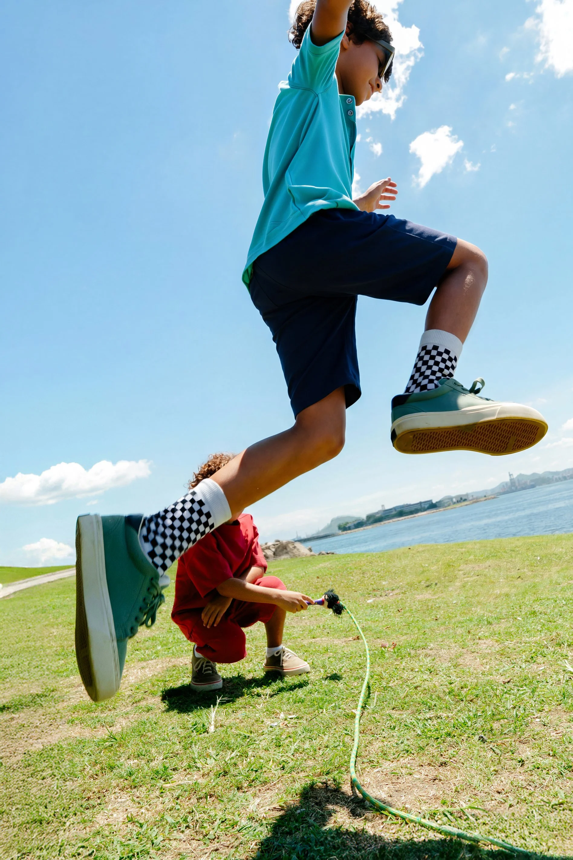 A young child and an older boy playing outdoors near a water body, with the boy jumping over the child. The children are wearing casual clothes, and the scene is sunny with blue skies.