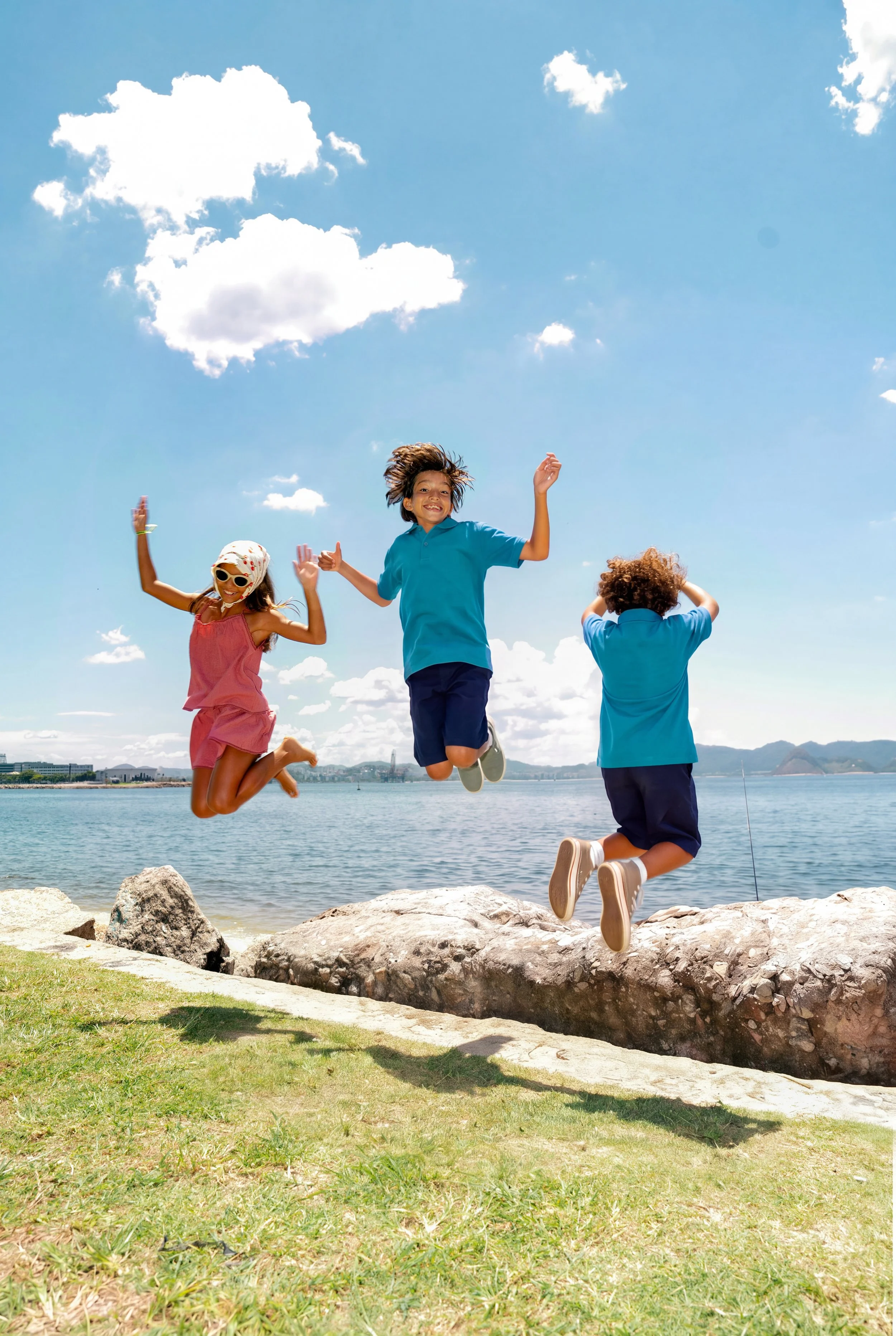 Three children jumping on rocks near a body of water under a partly cloudy sky, smiling and enjoying outdoor play.