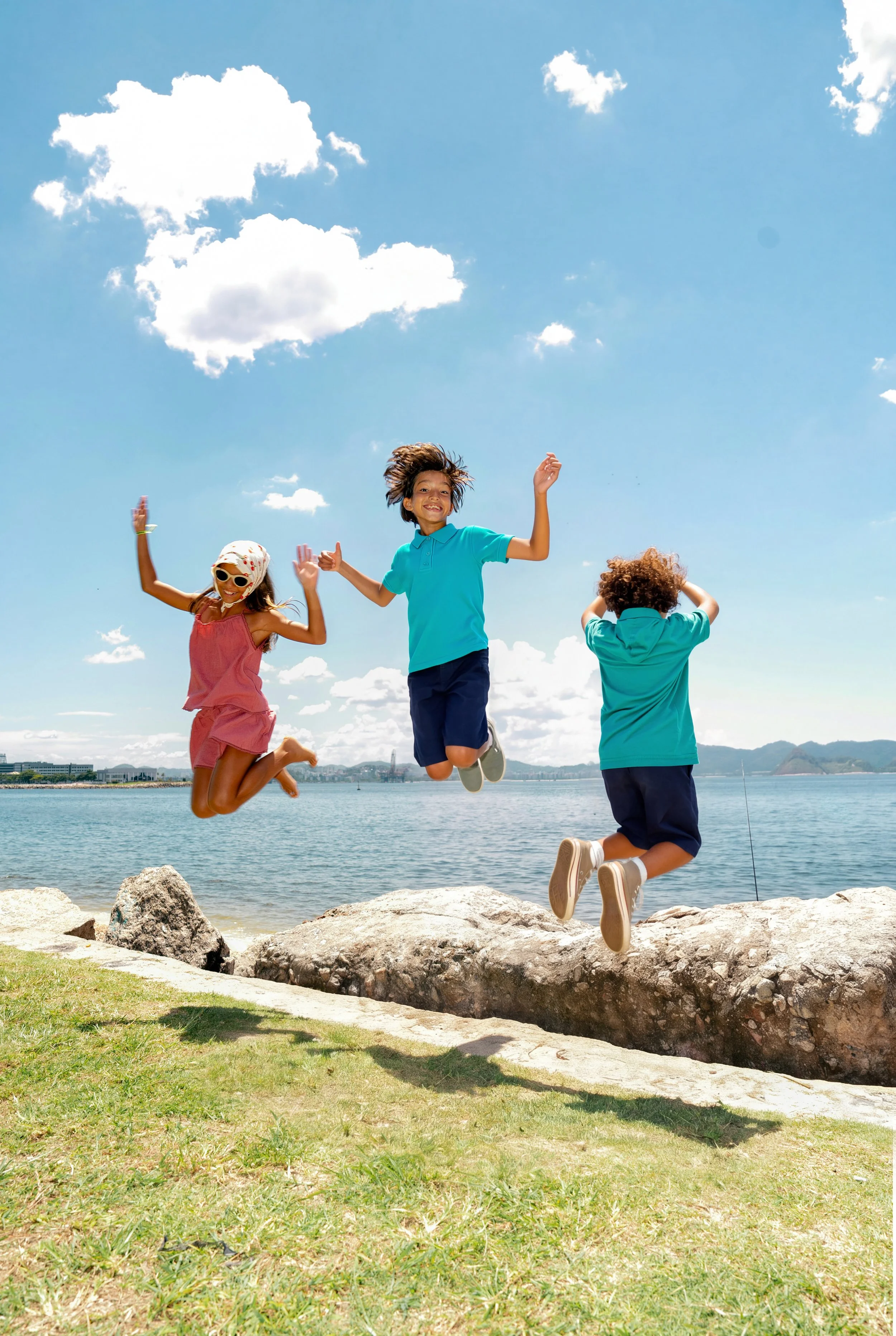 Three children jumping in the air by a lake on a sunny day. One girl wearing sunglasses, a white hat, and a pink dress; a boy in a blue shirt and shorts; another boy with curly hair in a turquoise shirt. Rocks and grass in the foreground, cityscape and mountains in the background.