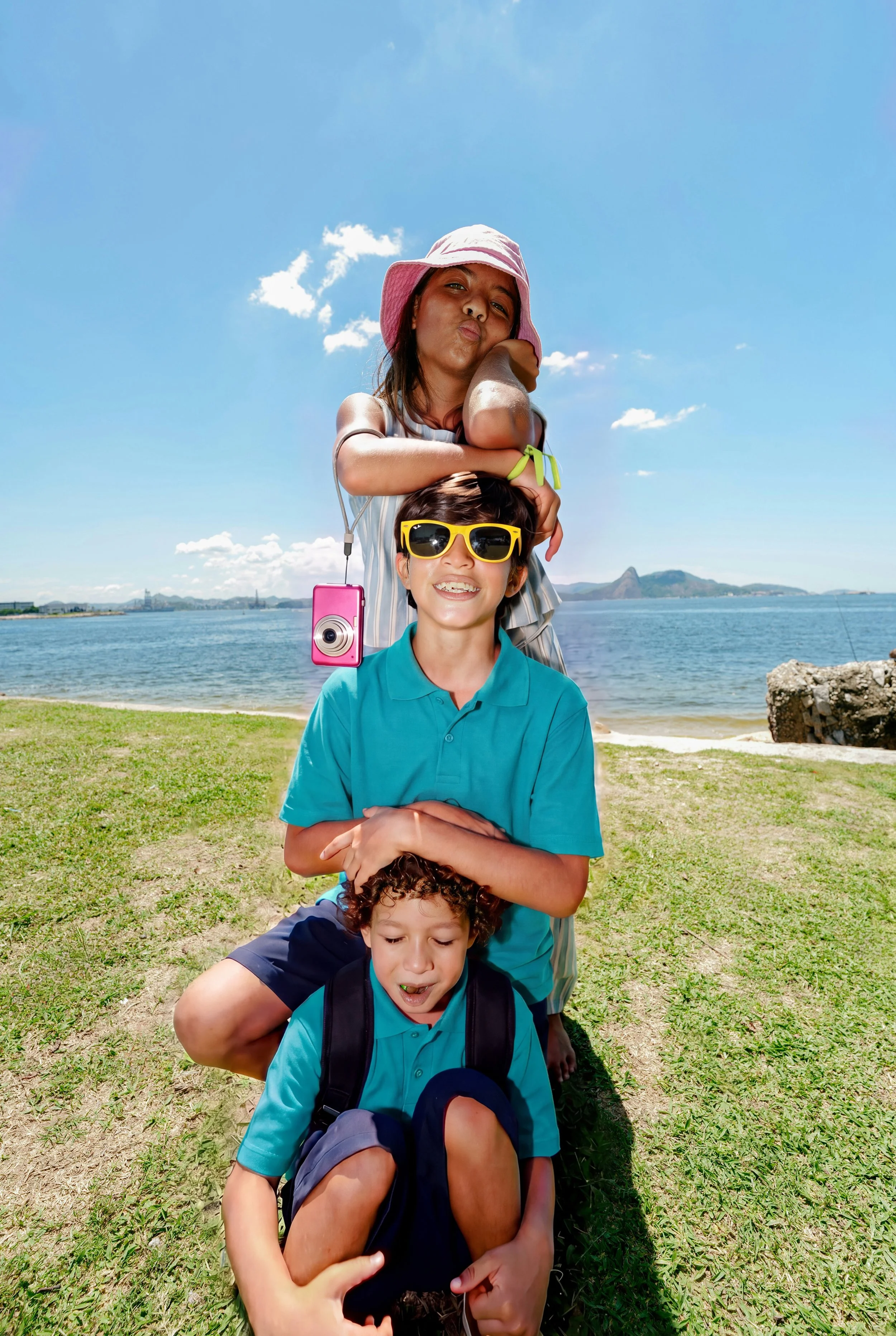 Five kids sitting in a line outdoors near a body of water, with blue sky and scattered clouds above. The kids are stacked, one on top of another, holding hands or resting on each other's shoulders, smiling and having fun.