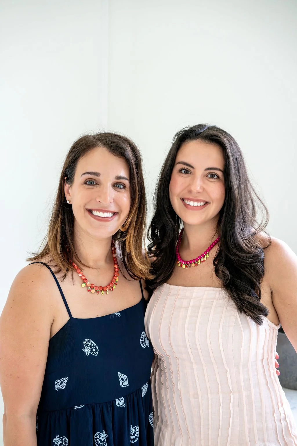 Two smiling women in summer dresses standing close together indoors, with a blank wall behind them.