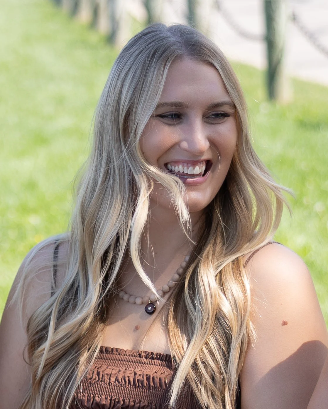 A young woman with long blonde hair smiling and looking to her left, outdoors in a grassy area.