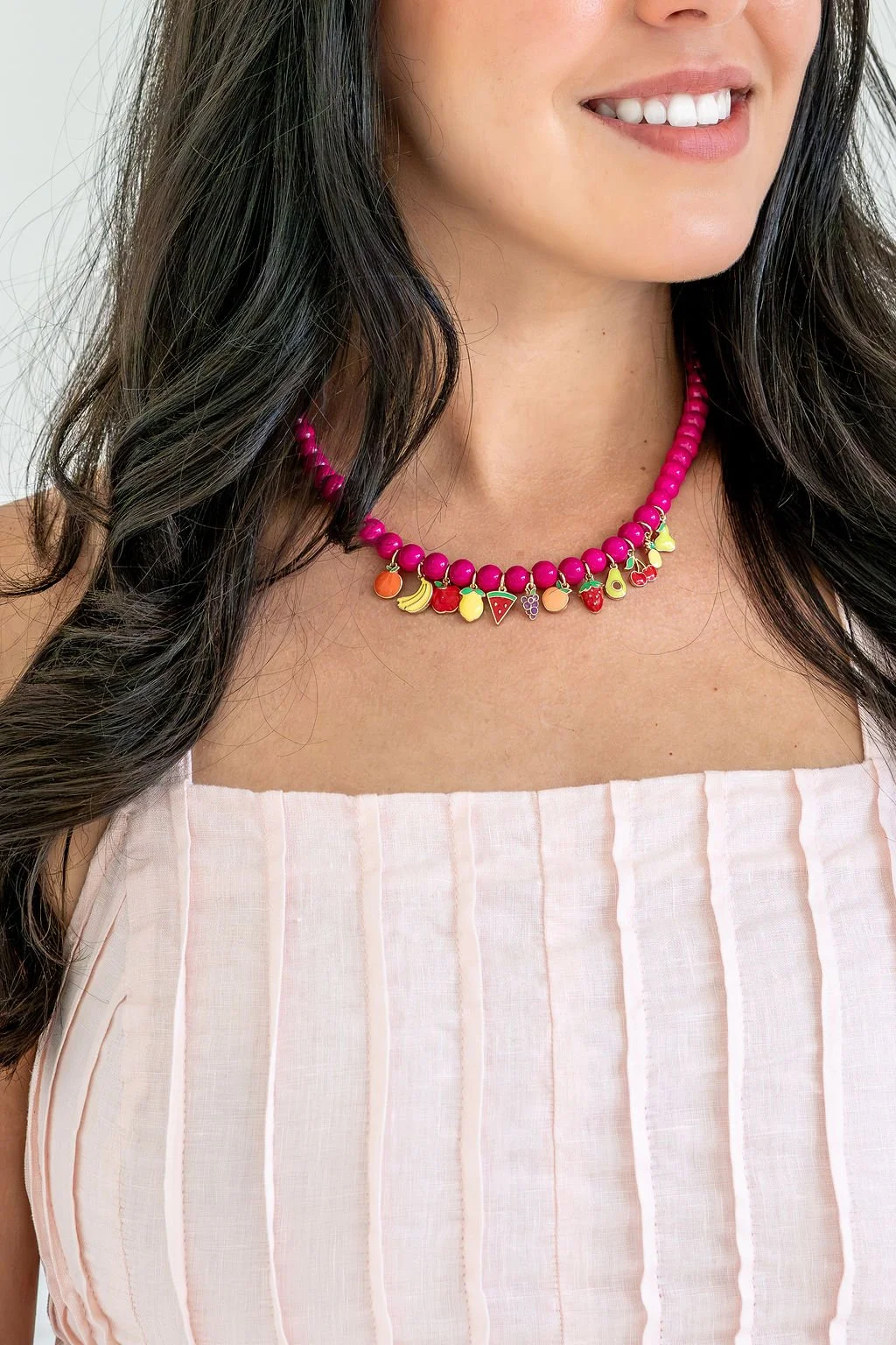 Close-up of a woman wearing a colorful fruit-themed bead necklace, smiling, with long dark hair and a white striped sleeveless top.