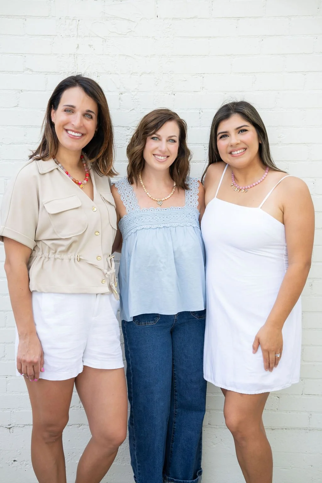 Three women standing closely together in front of a white brick wall, smiling at the camera. They are dressed casually, with the woman in the middle wearing a light blue top and jeans, and the women on either side wearing white dresses.