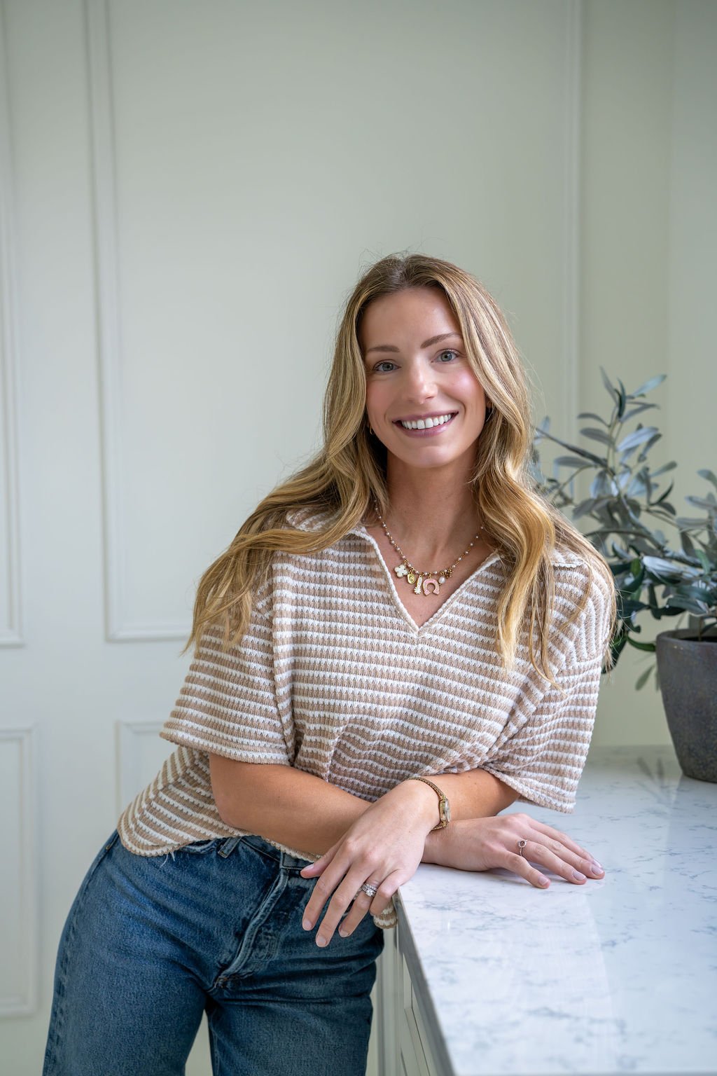 A woman with wavy blonde hair smiling, wearing a beige and white striped short-sleeve top, blue jeans, and jewelry, standing in a room with light-colored walls and a potted plant.