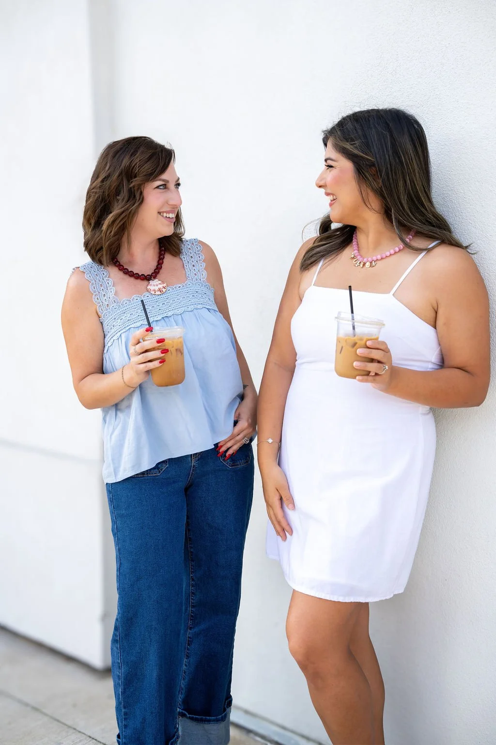 Two women stand against a white wall, smiling and holding iced coffee drinks, enjoying a conversation.