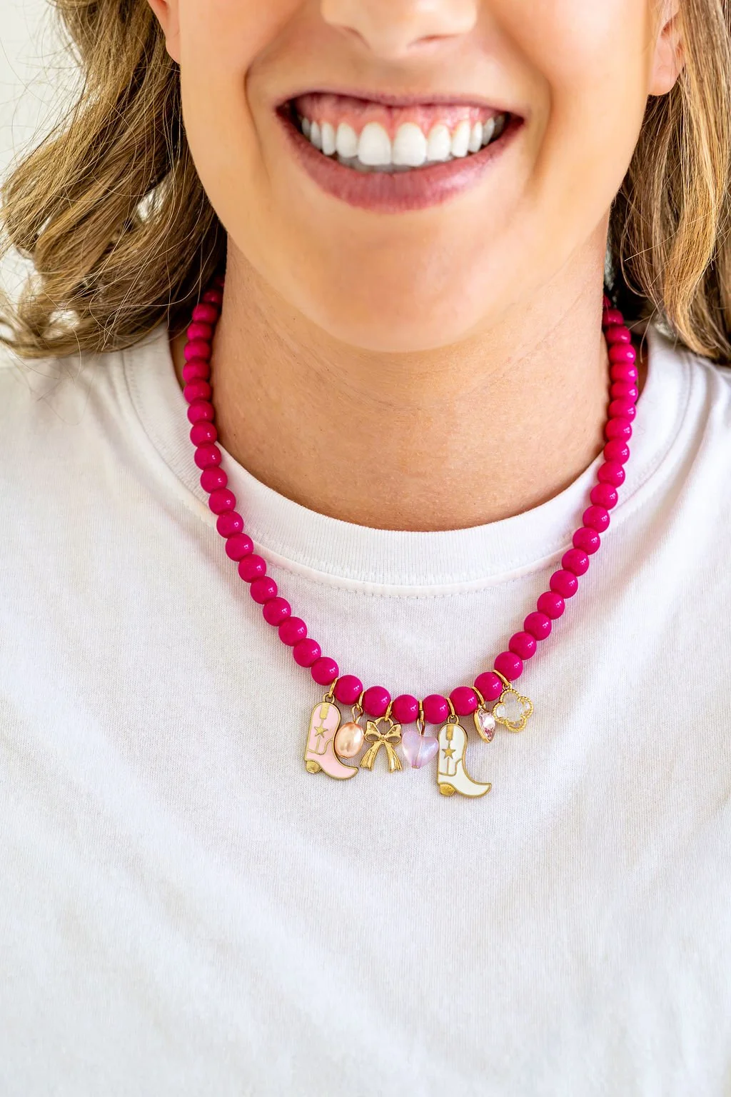 Close-up of a woman smiling, wearing a pink beaded necklace with charms, white shirt, and visible shoulder-length hair.